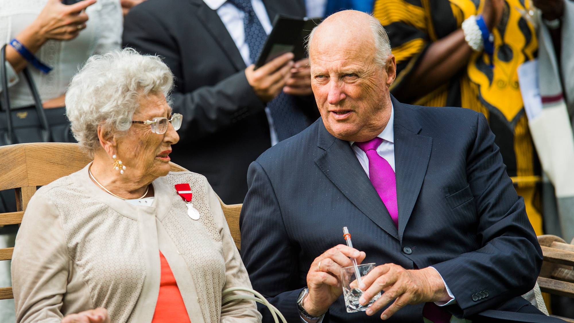 Kong Harald og en eldre kvinne sitter sammen på en benk. (Bildebeskrivelsen er laget av en KI-tjeneste)