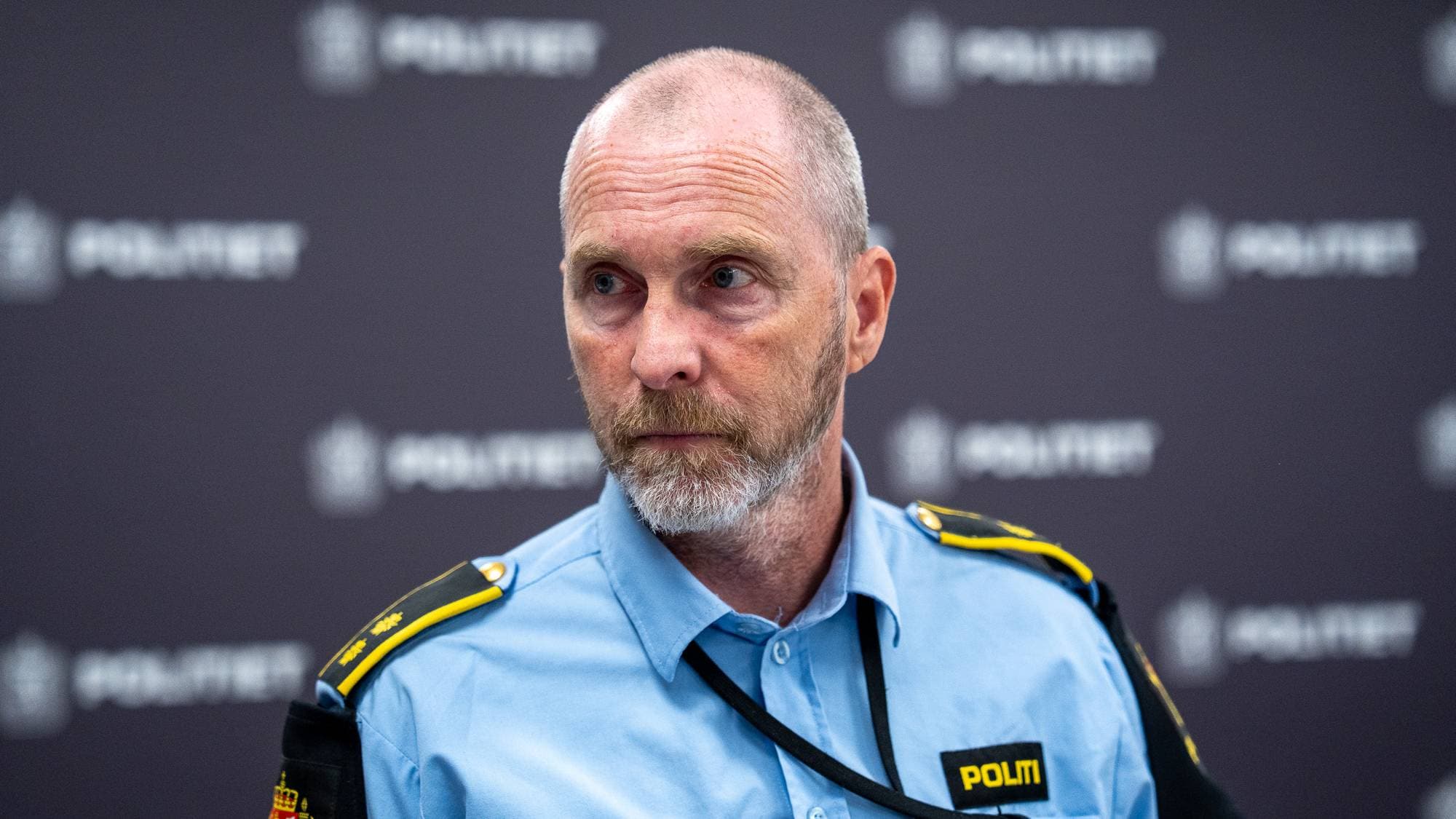 En mann i uniform fra politiet. Han har gråsprengt hår, skjegg og ser til siden. Bak ham er det et mørkt bakteppe med gjentatte "POLITIET"-logoer. (Bildebeskrivelsen er laget av en KI-tjeneste)