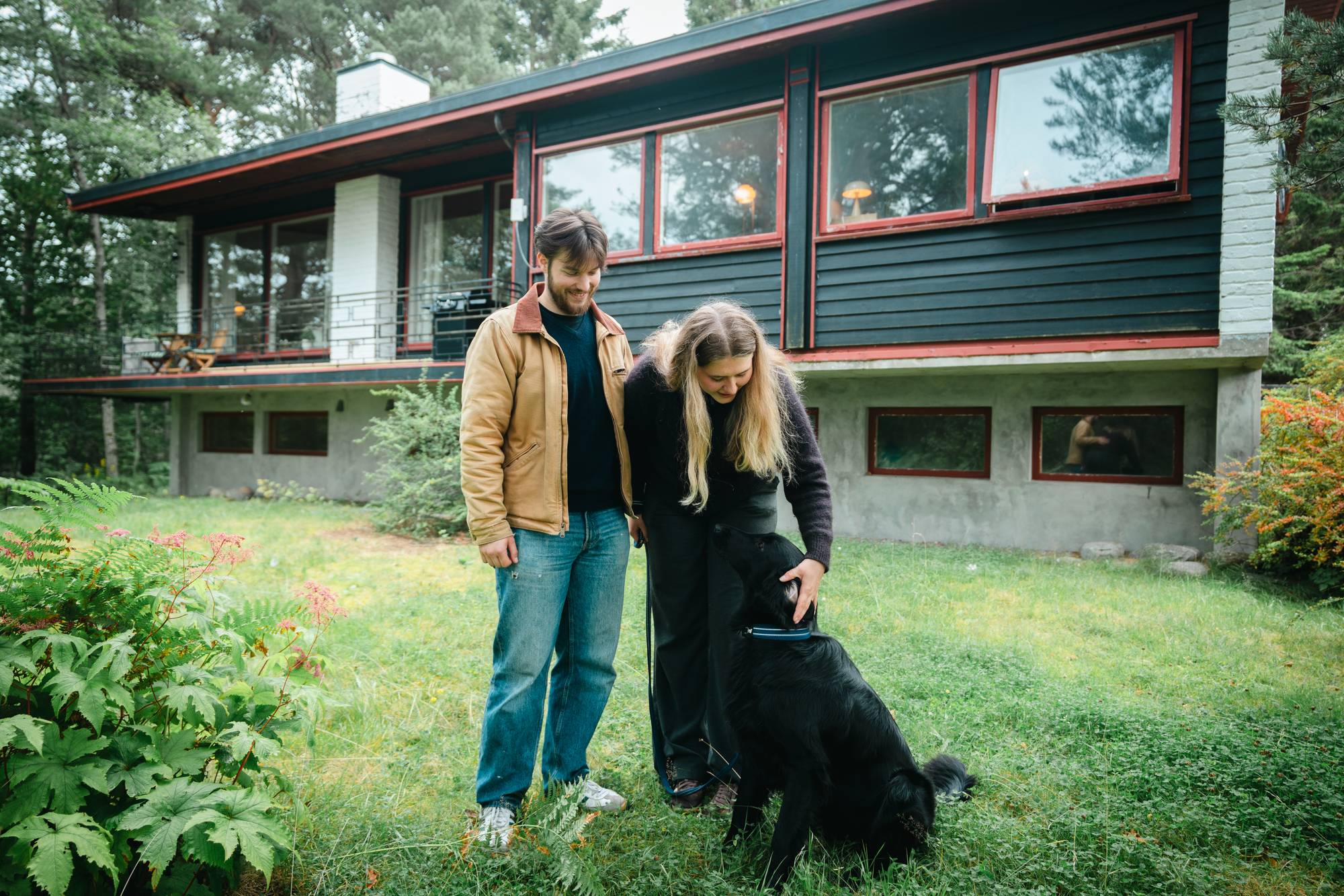 En mann og en kvinne står sammen med en svart hund i en hage foran et hus. (Bildebeskrivelsen er laget av en KI-tjeneste)