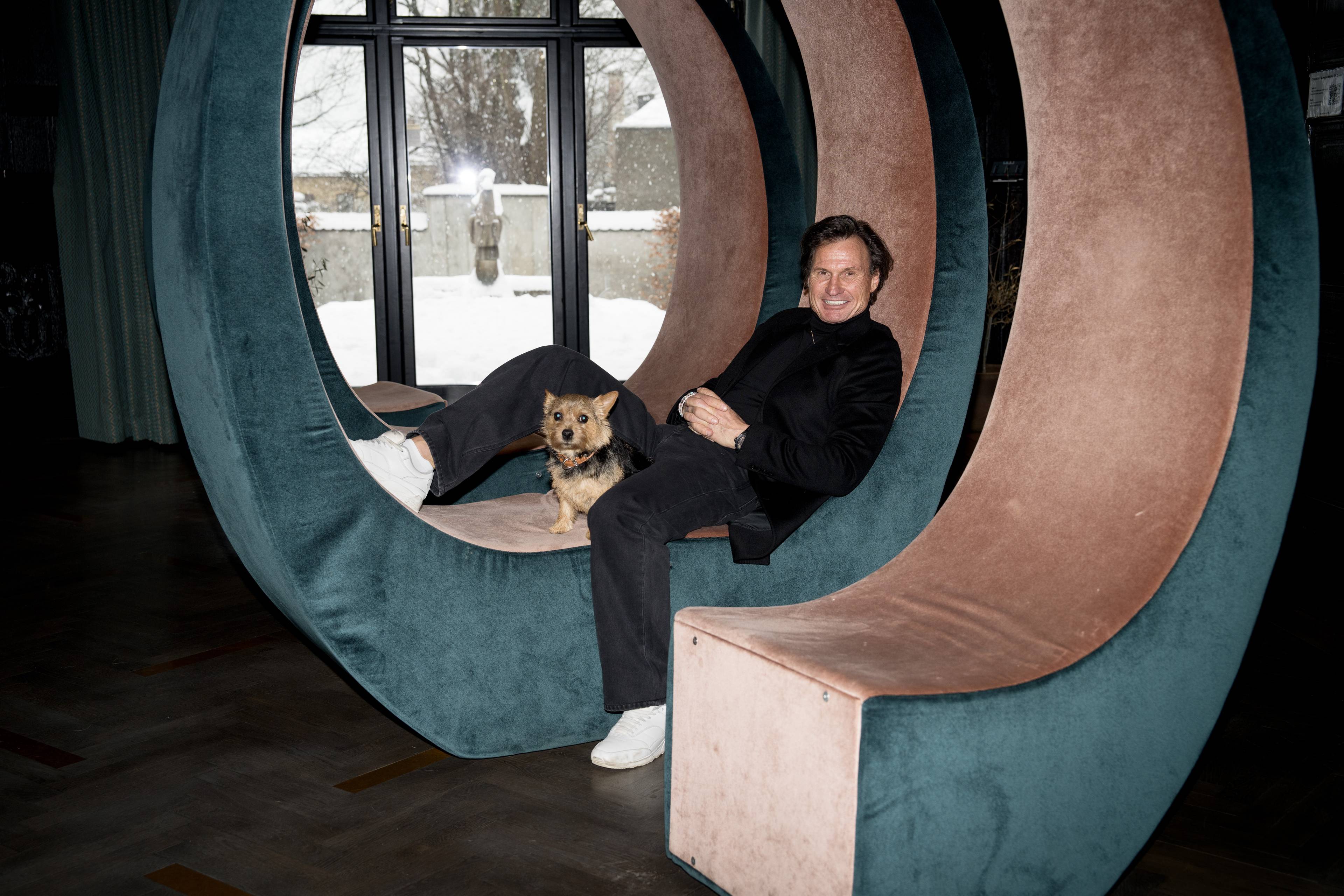 Petter Stordalen sitter i en spiralformet sofa, lener seg tilbake og har en hund på fanget.