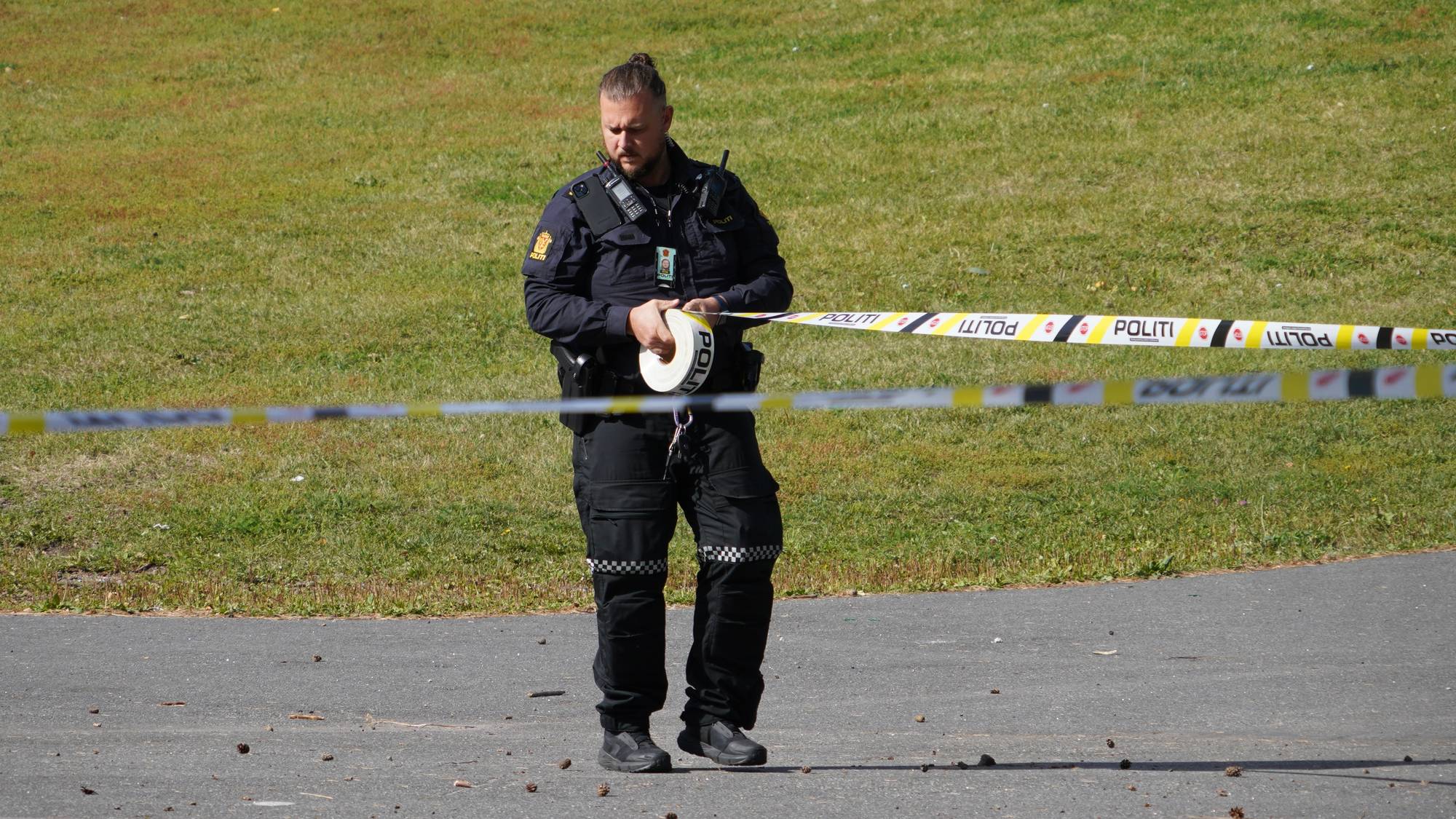 En politibetjent går og setter opp sperrebånd på et område med grønt gress i bakgrunnen