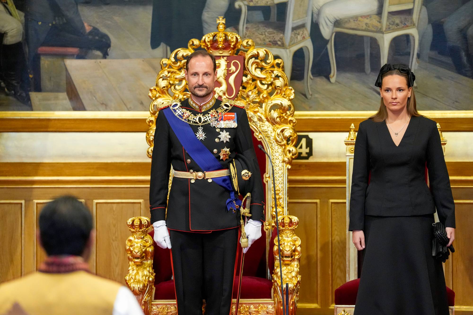 Kong Haakon og kronprinsesse Ingrid Alexandra står foran kongestolen og kronprinsessestolen i stortingssalen under den høytidelige åpningen av det 171. storting. De ser mot stortingspresident Masud Gharahkhani, som står foran dem.
