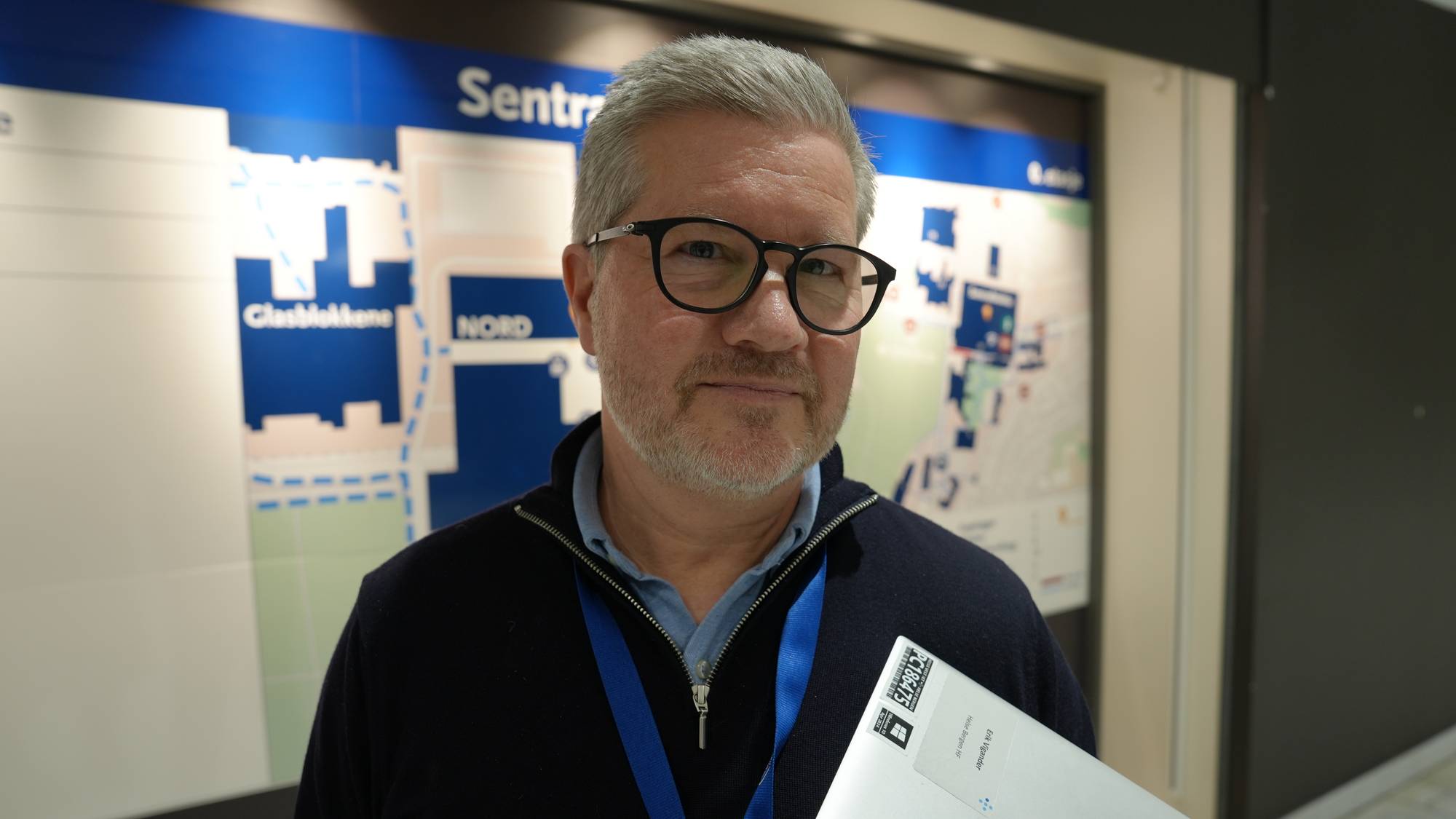 En person står foran et informasjonsskilt i en innendørs setting. Personen holder en mappe og er kledd i en blå genser. Skiltet bak viser en oversikt over området med ulike ikoner og stier. Opplysningene indikerer at dette kan være en offentlig plass eller et kjøpesenter.