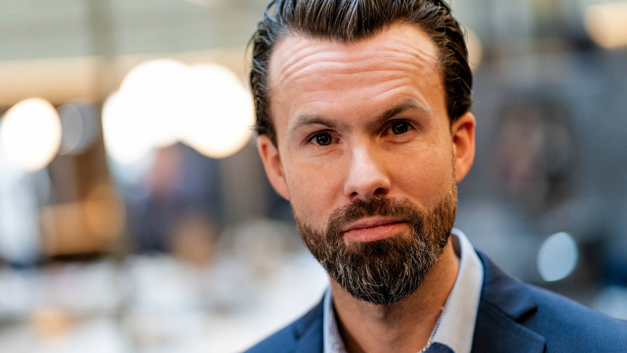 Et portrett av en mann med mørkt, kort hår og skjegg, iført en blå dressjakke og lys skjorte. Han står i en moderne innendørs setting med dempet belysning og uskarpe bakgrunner. Bakgrunnen viser flere uskarpe figurer som kan være mennesker eller objekter. Bildet gir inntrykk av en profesjonell atmosfære. (Bildebeskrivelsen er laget av en KI-tjeneste)