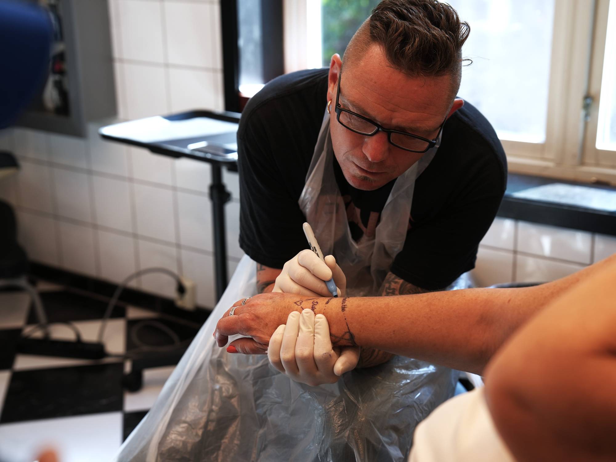 Remi har på seg briller og tegner på hånden til en kvinne som skal tatoveres