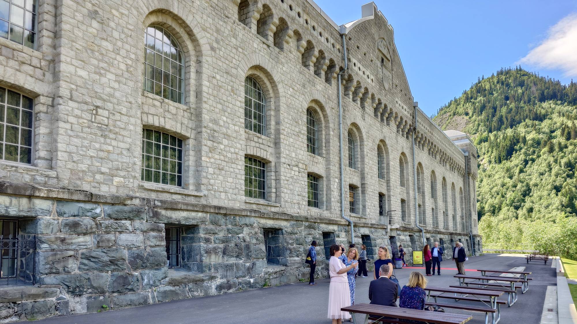 Personer oppholder seg utenfor den store steinbygningen Vemork på Rjukan. (Bildebeskrivelsen er laget av en KI-tjeneste)