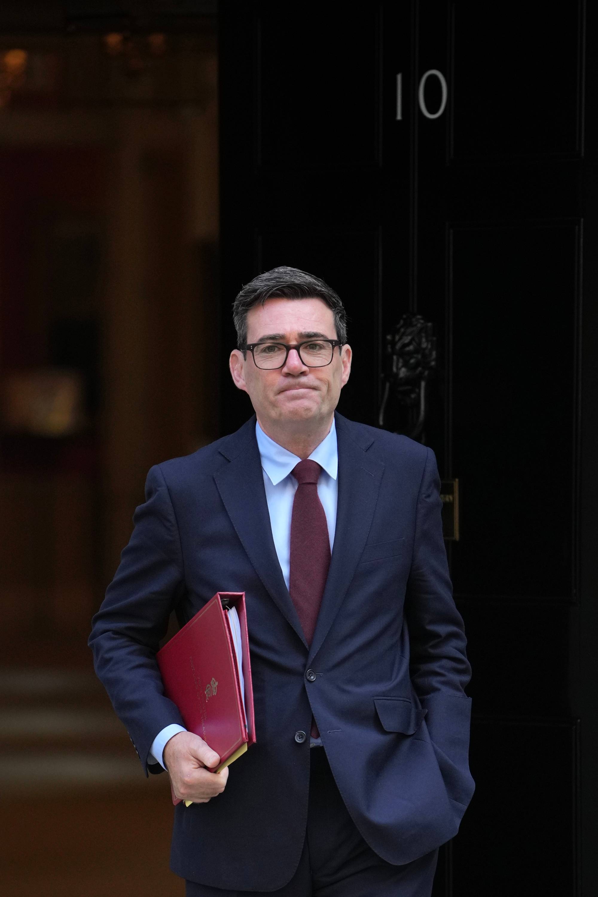 Andy Burnham i dress og briller foran inngangen til 10 Downing Street med en rød mappe i hånden. (Bildebeskrivelsen er laget av en KI-tjeneste)