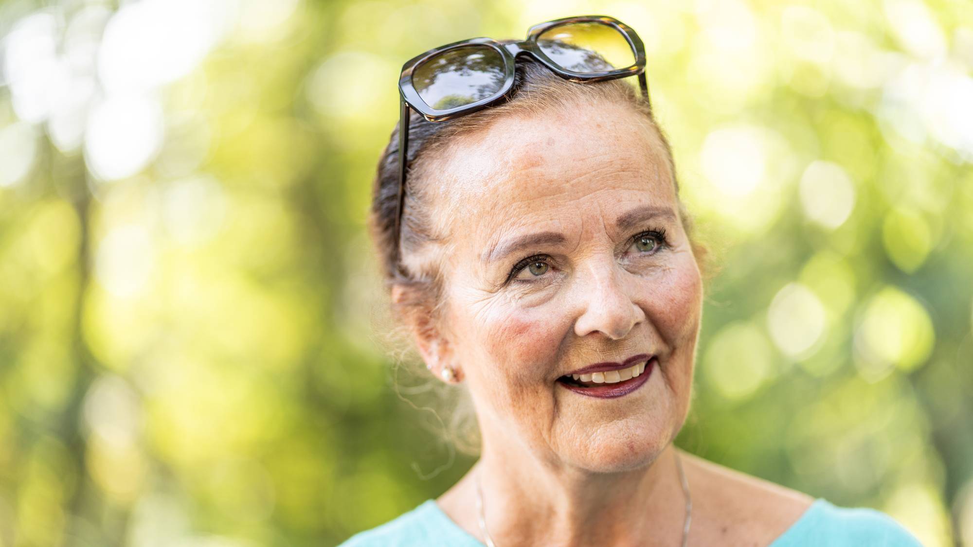 Portrett av en smilende kvinne i lyseblå topp med solbriller i håret utendørs. (Bildebeskrivelsen er laget av en KI-tjeneste)