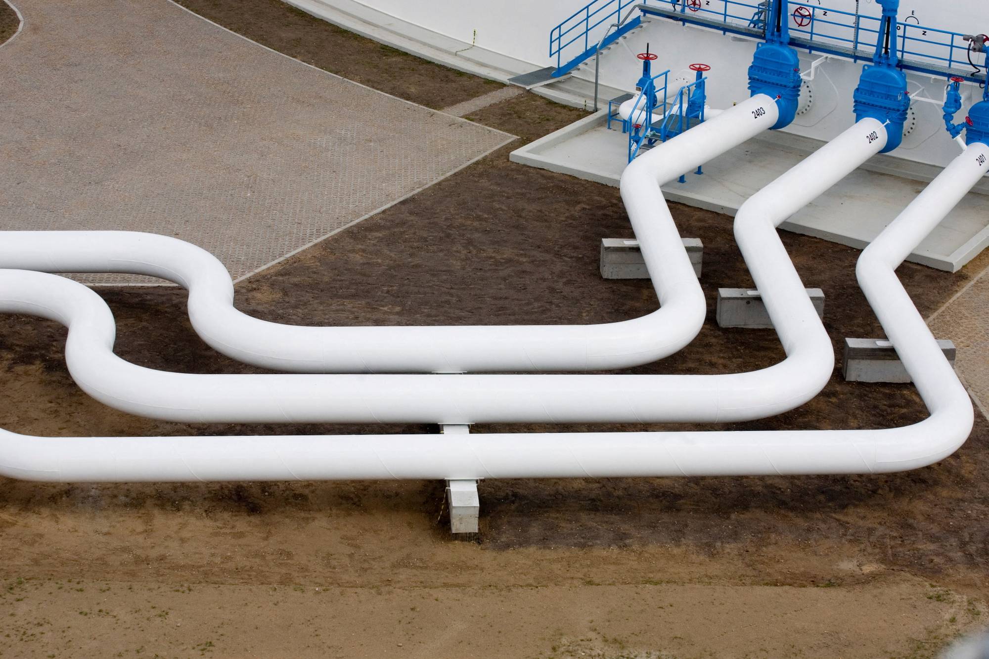 Et industriområde med store hvite rør som går over bakken. Rørene er koblet til en blå og hvit installasjon med ventiler. Bakken er dekket av grus og belegningsstein. (Bildebeskrivelsen er laget av en KI-tjeneste)