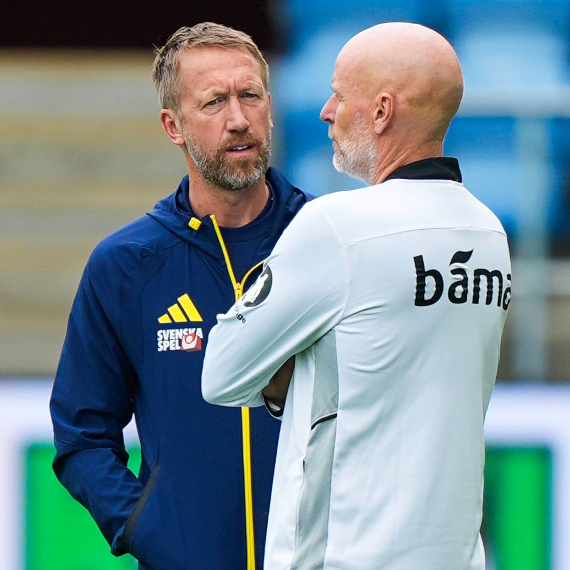 To fotballtrenere, Graham Potter og en ukjent mann, diskuterer på en bane. Potter, iført en mørkeblå jakke med sponsorlogoer, snakker med den andre mannen. Han har grått skjegg og ser fokusert ut. Mannen ved siden av ham er skallet og har på seg en hvit treningsgenser med en stor logo på ryggen. (Bildebeskrivelsen er laget av en KI-tjeneste)