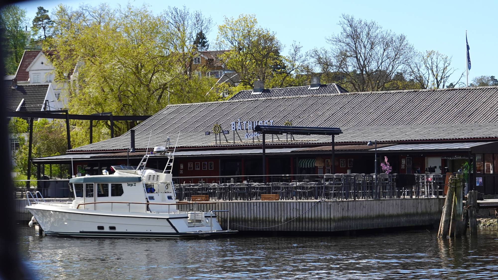 En hvit motorbåt ligger fortøyd ved en restaurant med uteservering. Restauranten har et bredt tak og mange bord og stoler. I bakgrunnen ses flere hus og trær. (Bildebeskrivelsen er laget av en KI-tjeneste)