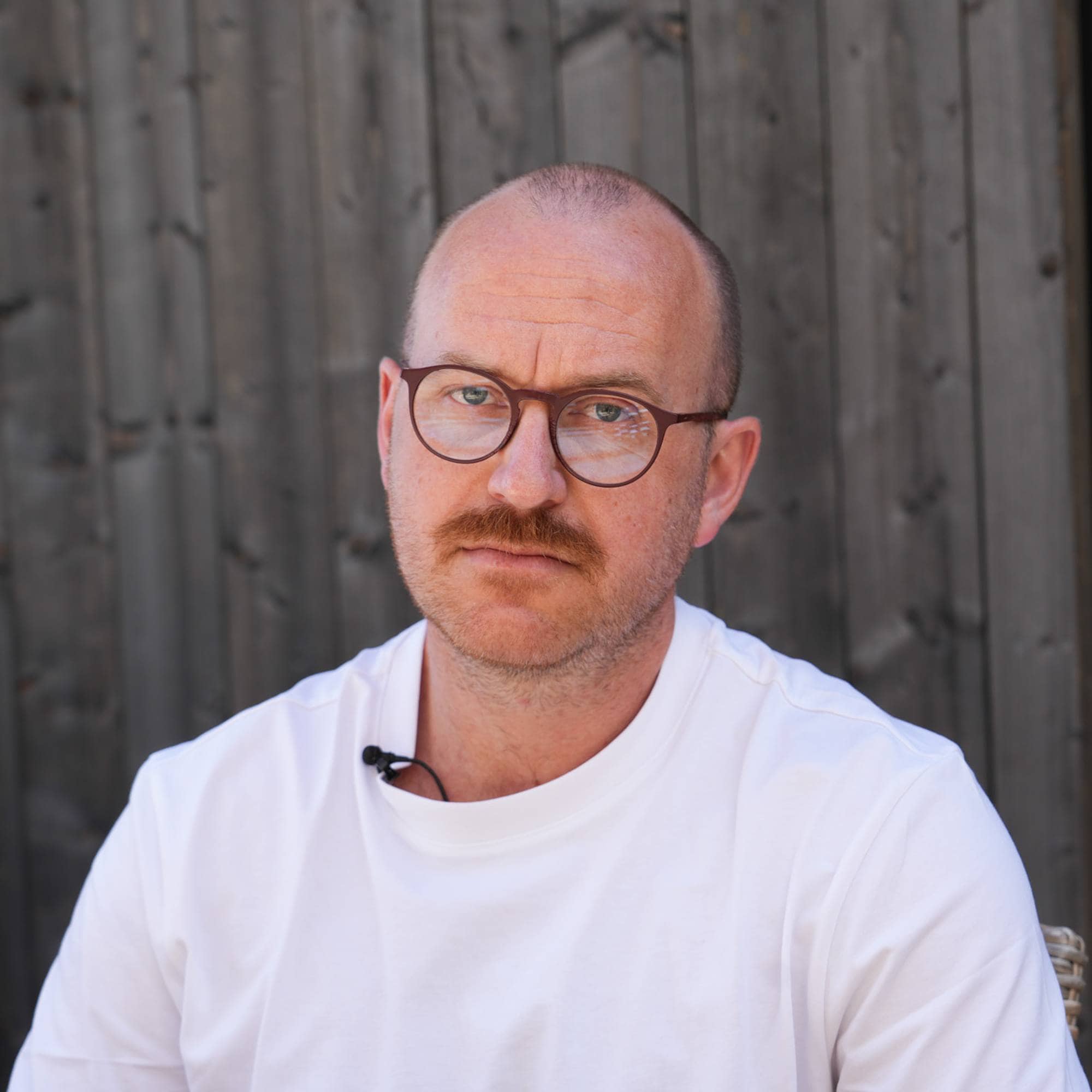 Portrett av en mann som sitter ute. Han har på en hvit t-skjorte og briller. 