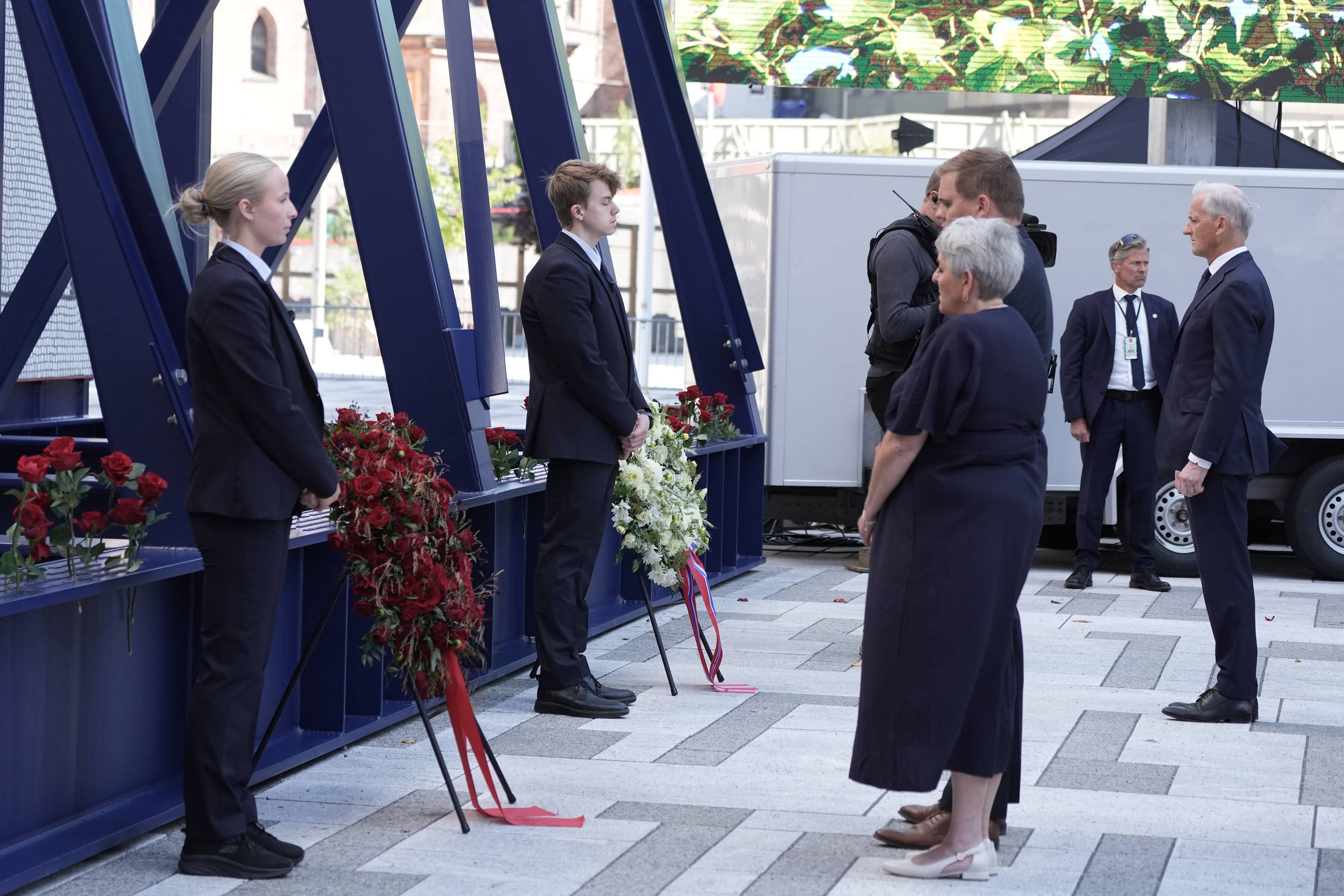 Statsminister Jonas Gahr Støre og andre står ved blomster ved regjeringskvartalet i Oslo. (Bildebeskrivelsen er laget av en KI-tjeneste)