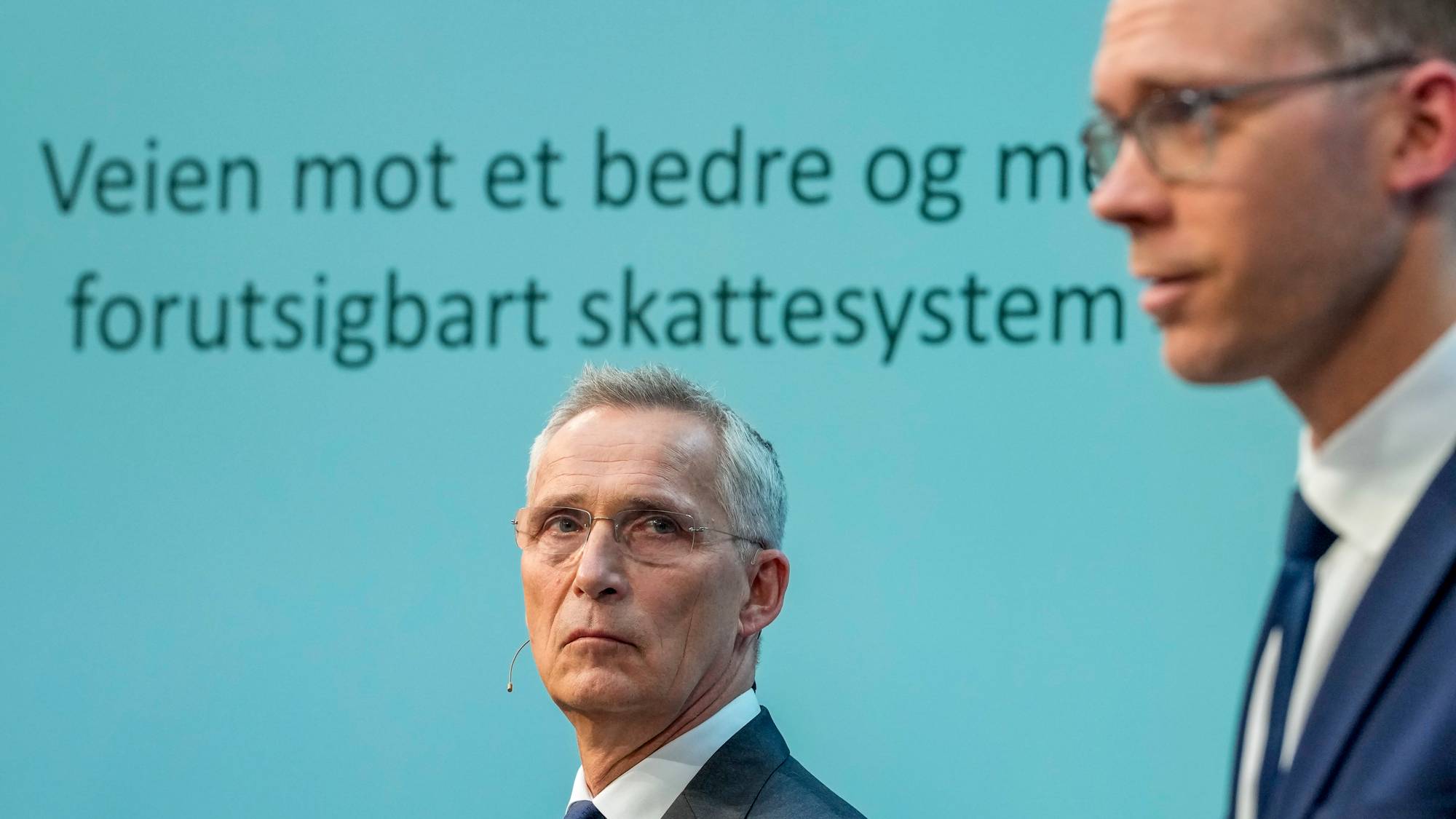 To menn står foran en lys blå skjerm. På skjermen står teksten "Veien mot et bedre og mer forutsigbart skattesystem". Den ene mannen, iført dress og slips, ser mot venstre. Den andre mannen, også i dress og slips, ser til siden. (Bildebeskrivelsen er laget av en KI-tjeneste)