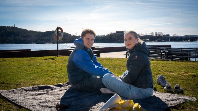 En ung mann og en ung kvinne sitter på et teppe på en gressplen ved vannet. De smiler mot kameraet. I bakgrunnen er en strandpromenade med benker og et livredningsutstyr. Solen skinner fra en klar, blå himmel. (Bildebeskrivelsen er laget av en KI-tjeneste)