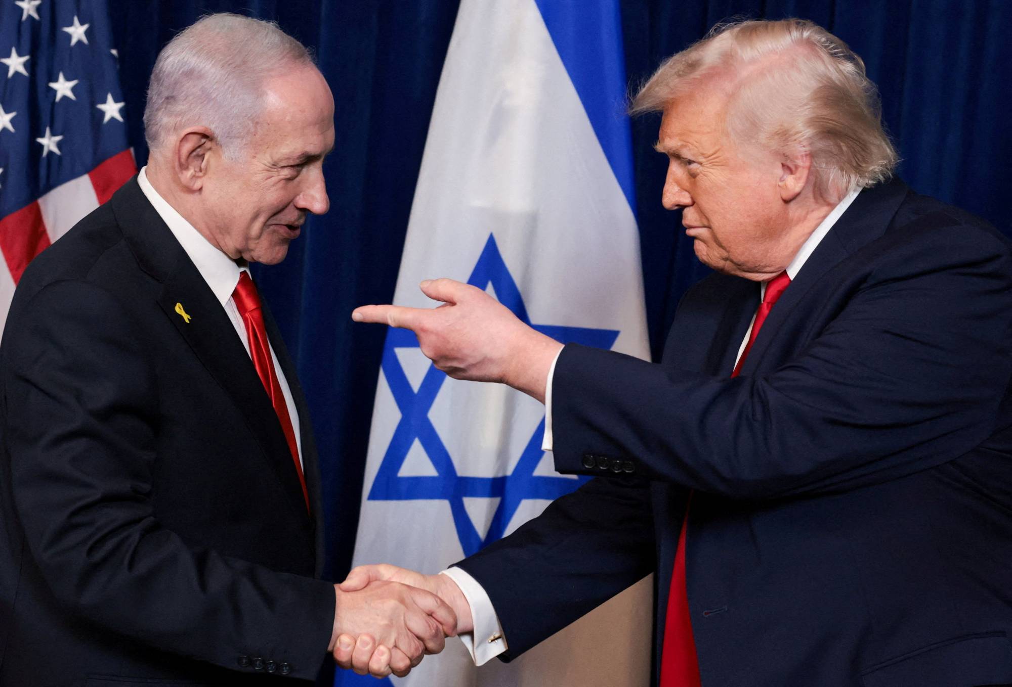 USAs president Donald Trump og Israels statsminister Benjamin Netanyahu håndhilser. Bak dem sees et amerikansk og et israelsk flagg. Netanyahu har på seg en gul sløyfe. (Bildebeskrivelsen er laget av en KI-tjeneste)