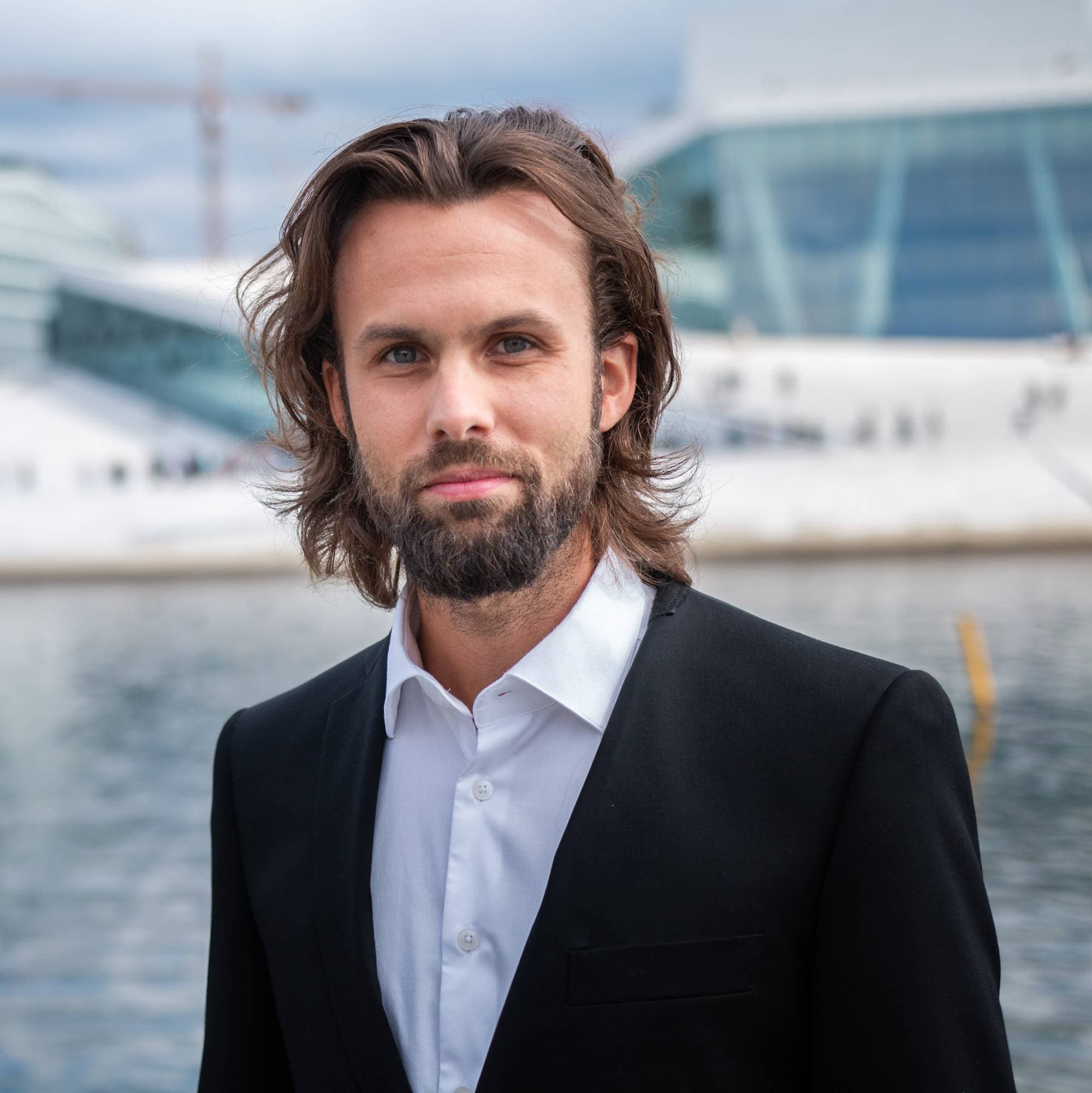 En mann med skjegg og mørkt hår som står foran Operaen og ser alvorlig ut i dress.