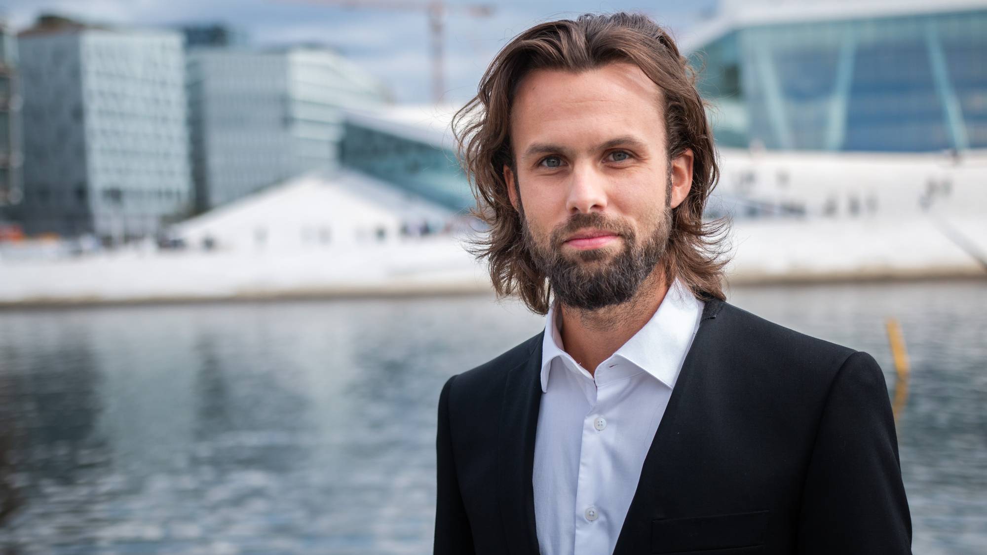 En mann med skjegg og mørkt hår som står foran Operaen og ser alvorlig ut i dress.