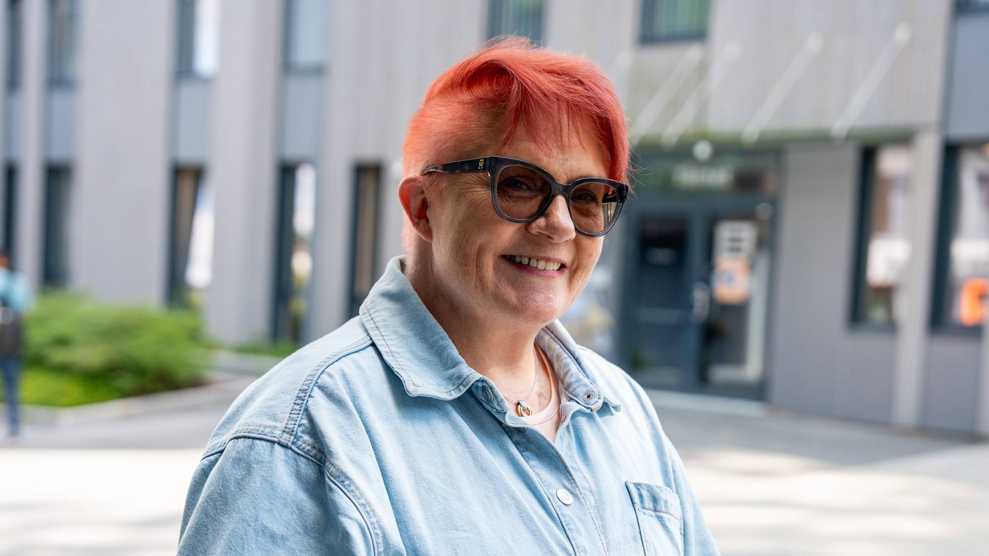 Kvinne med kort, rødt hår og briller, kledd i lyseblå denimskjorte, smiler utendørs.