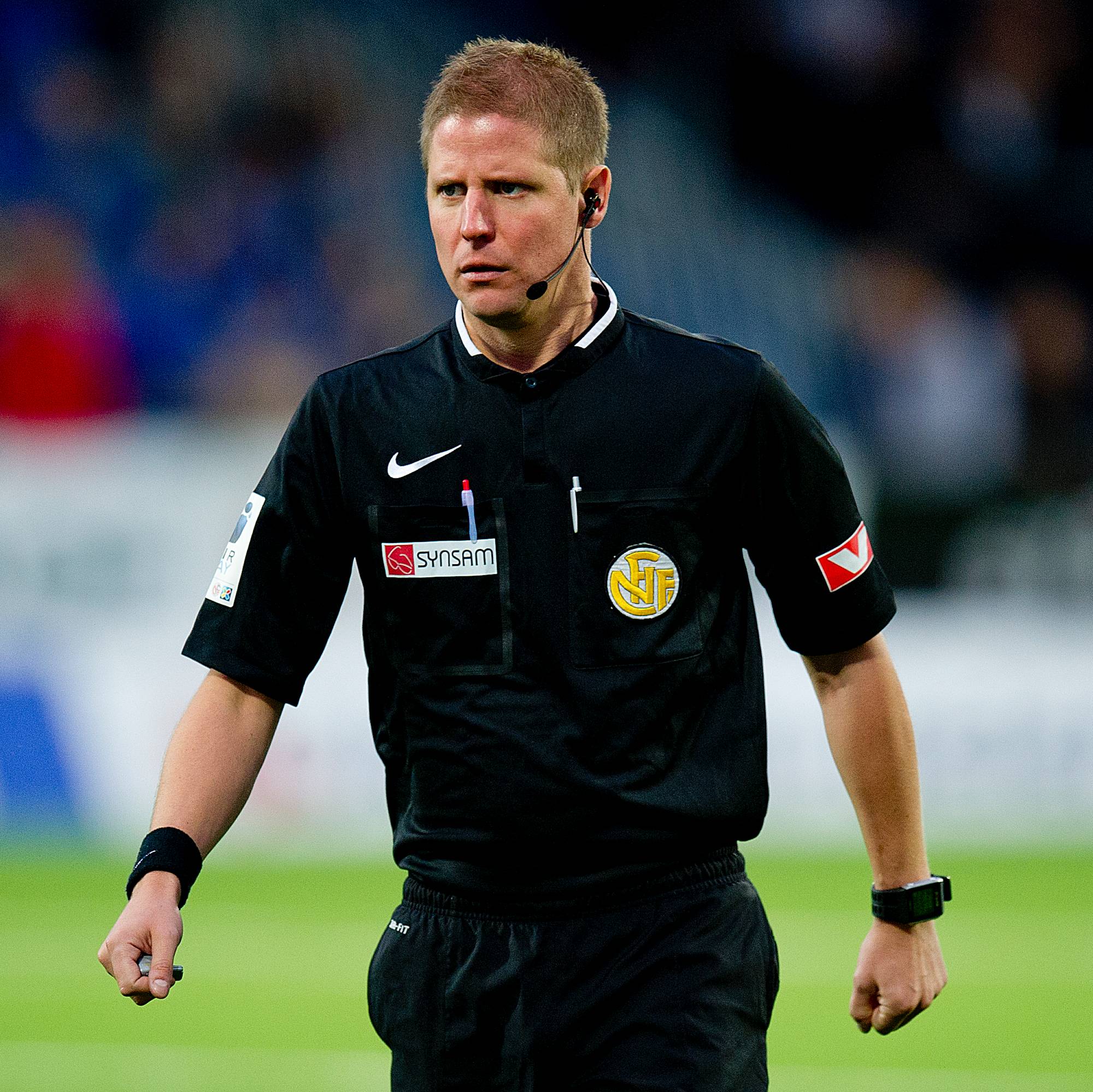 Fotballdommer i svart drakten går over gressmatta på en stadion. (Bildebeskrivelsen er laget av en KI-tjeneste)