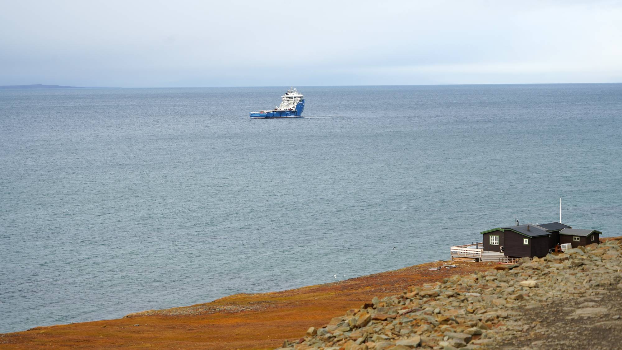 Et skip på havet. I forgrunn en hytte på land. 