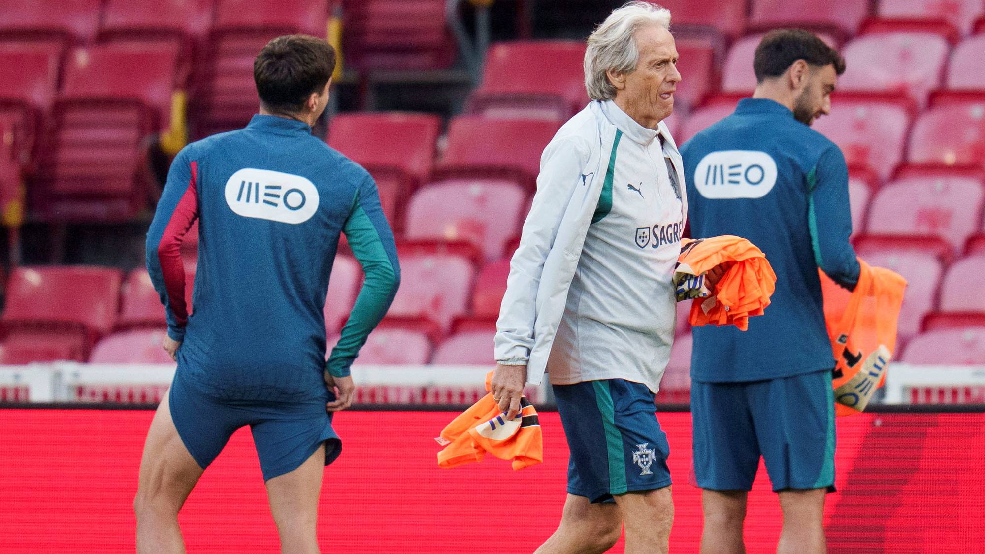 Trener Jorge Jesus går over en fotballbane med to spillere på en arena med røde seter. (Bildebeskrivelsen er laget av en KI-tjeneste)
