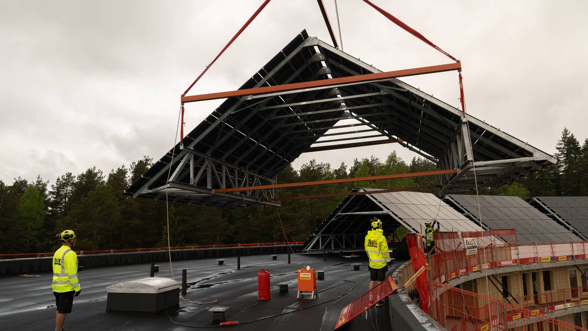 Et stort skråtak med solceller, som på et vanlig bolighus, heises på plass på taket på det store boligkomplekset. To bygningsarbeidere med gule jakker står ved siden av.