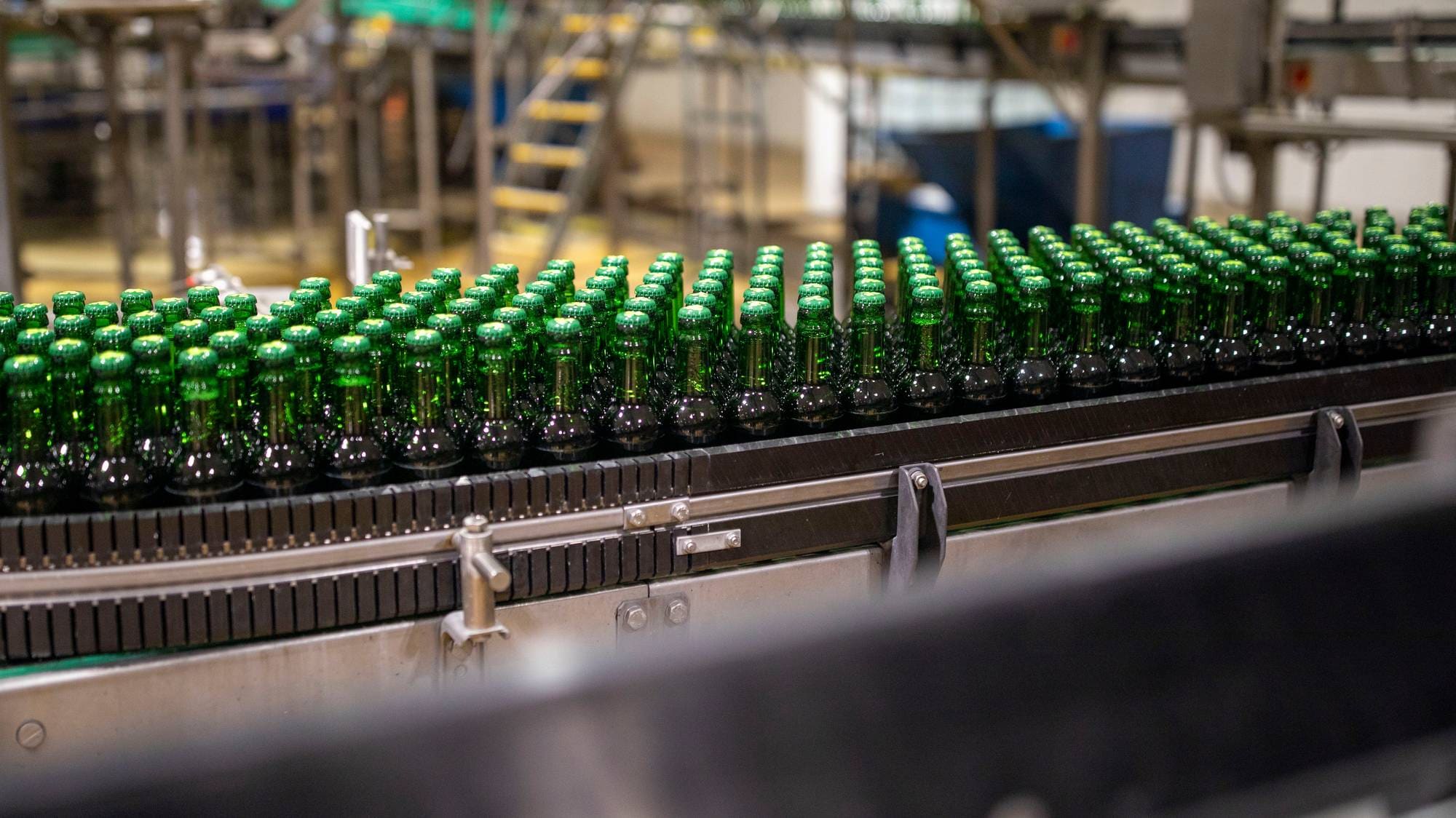 Mange grønne glassflasker på samlebånd i et produksjonslokale. (Bildebeskrivelsen er laget av en KI-tjeneste)
