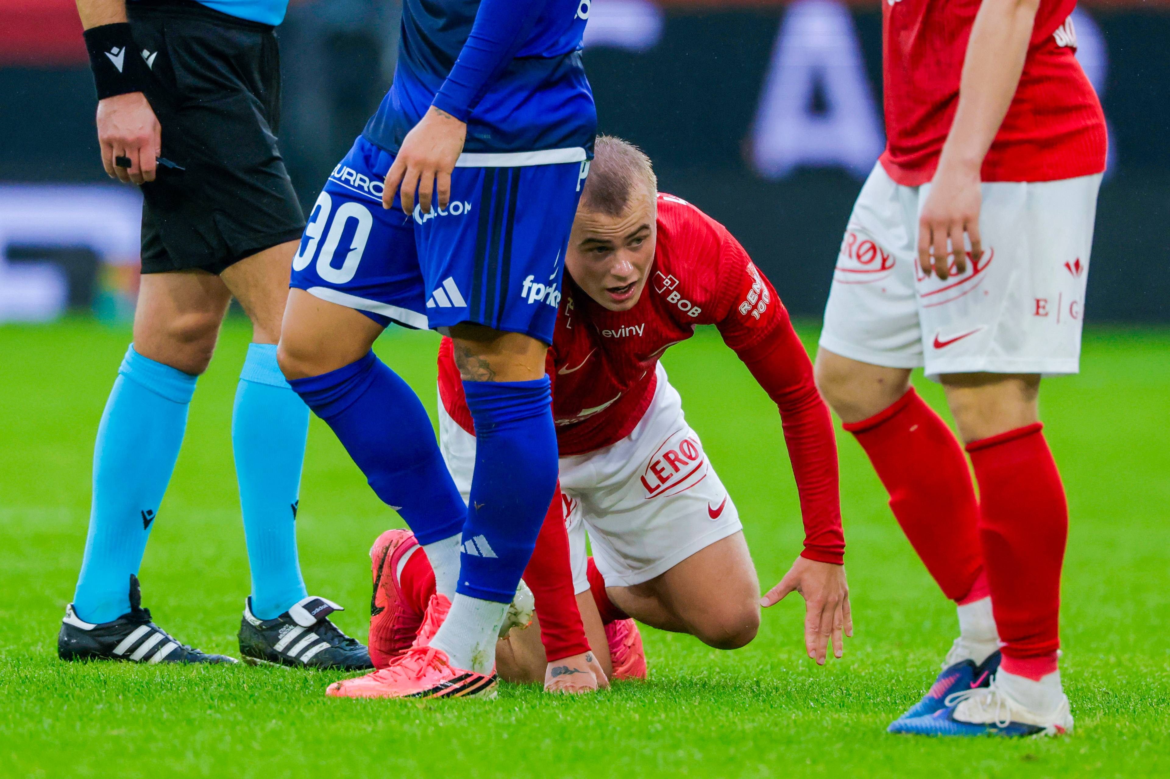 Fotballspiller i rød drakt sitter på banen omgitt av spillere i blått og rødt under en kamp. (Bildebeskrivelsen er laget av en KI-tjeneste)