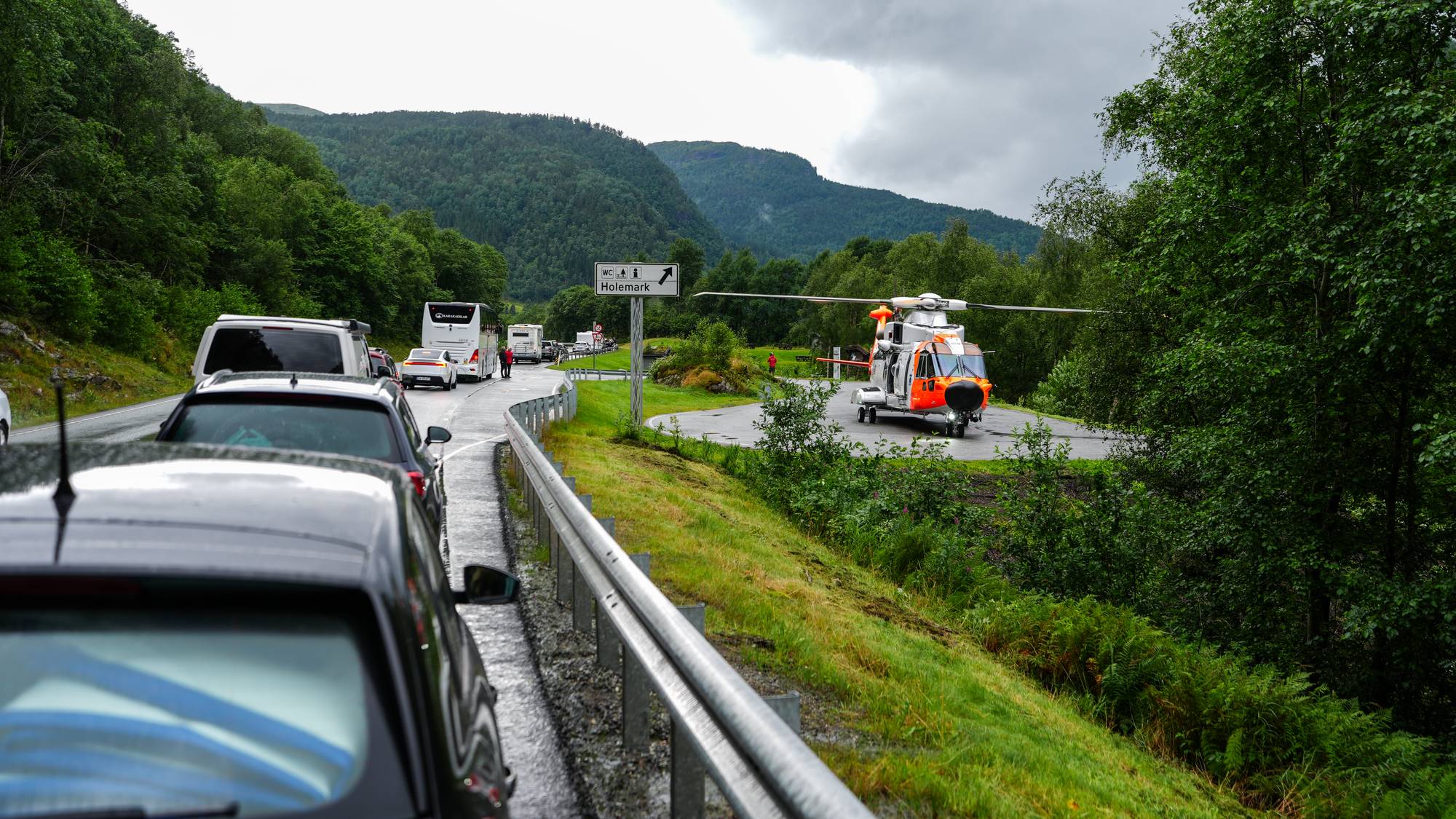 Redningshelikopter står på en rasteplass ved en kø på en vei i et skogkledd landskap. 