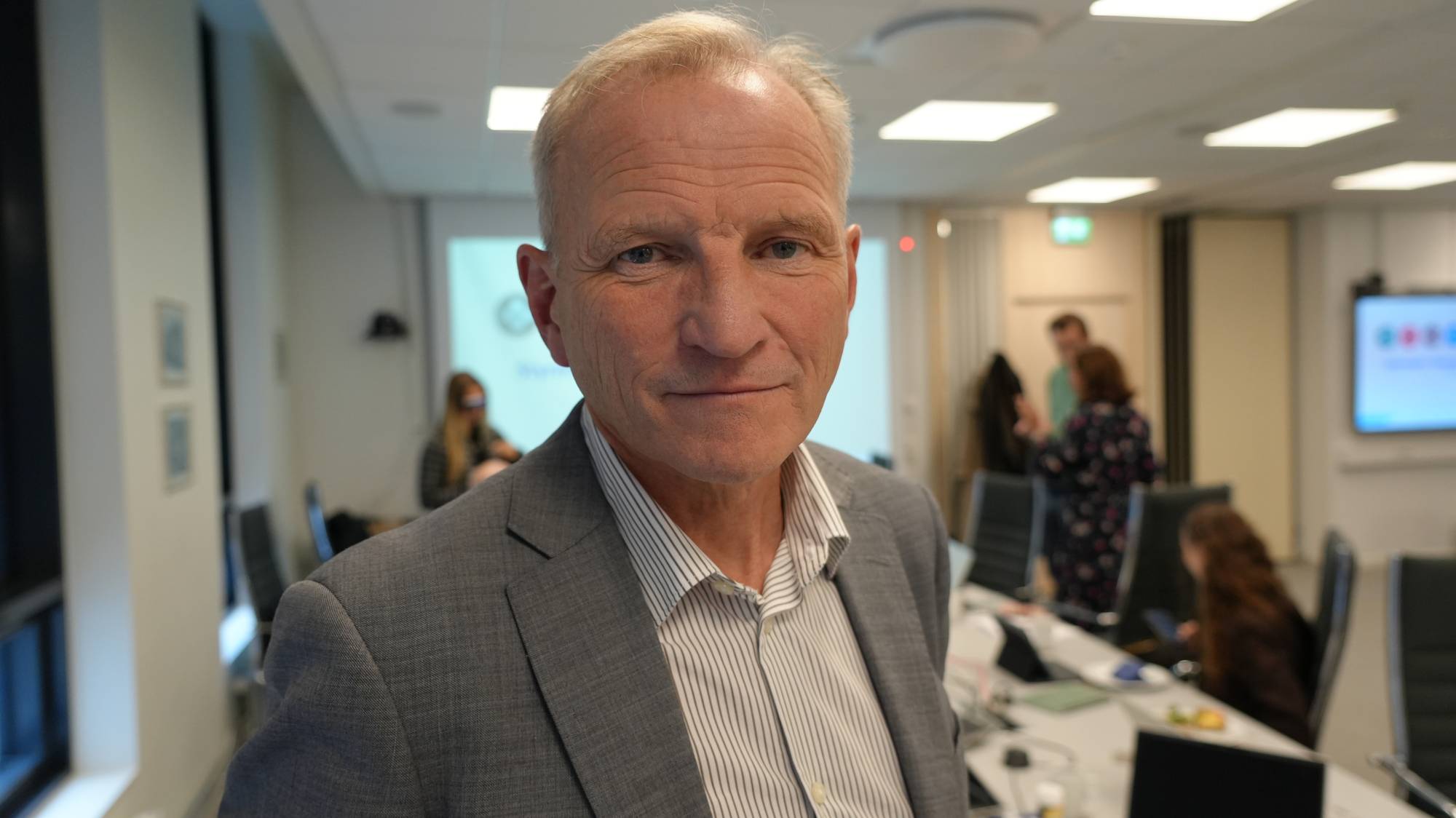 Geir Pollestad i en grå dress står foran et møterom med flere personer i bakgrunnen. (Bildebeskrivelsen er laget av en KI-tjeneste)
