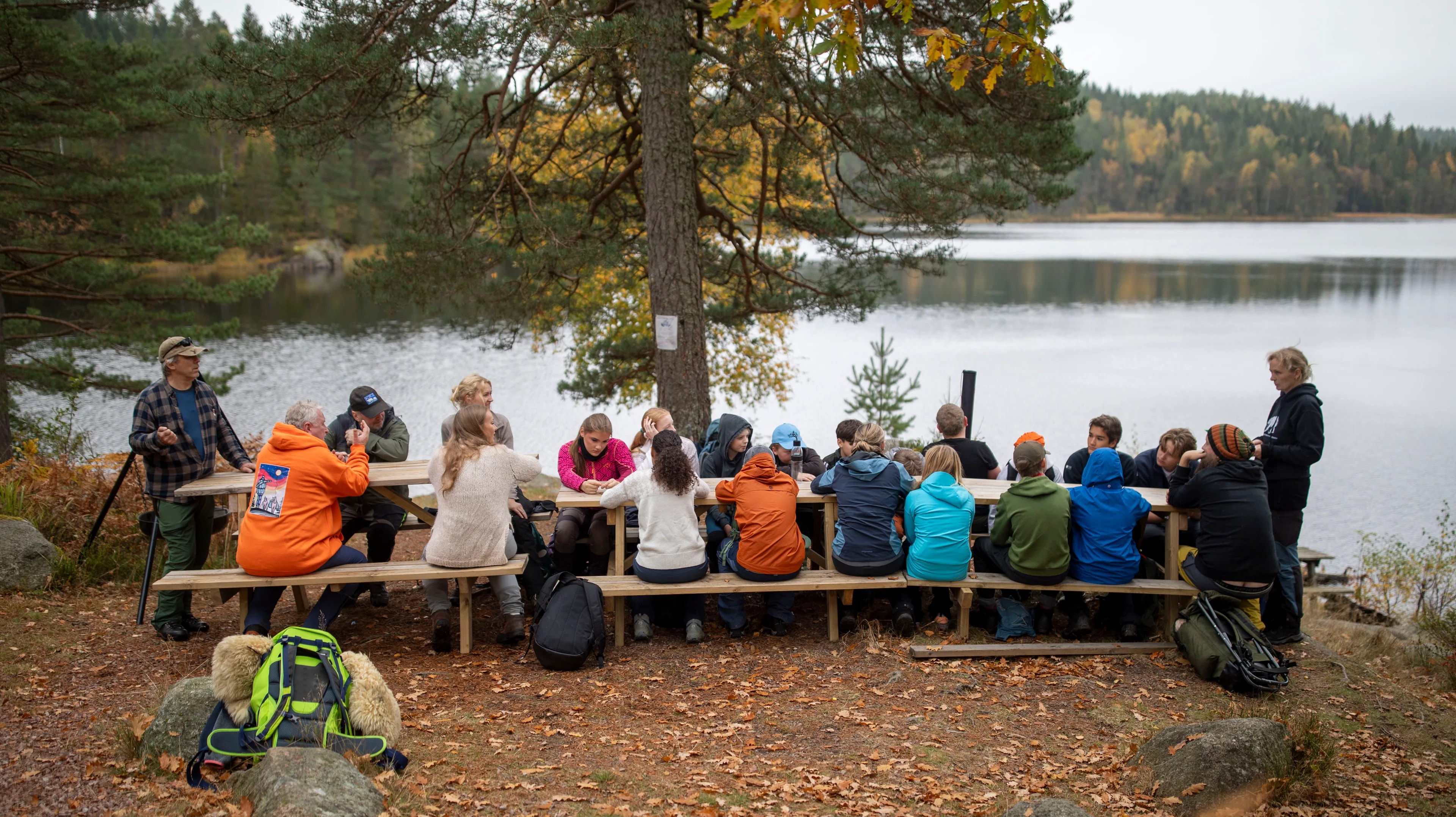 En stor gruppe mennesker sitter rundt et langbord ved et vann i skogen. (Bildebeskrivelsen er laget av en KI-tjeneste)