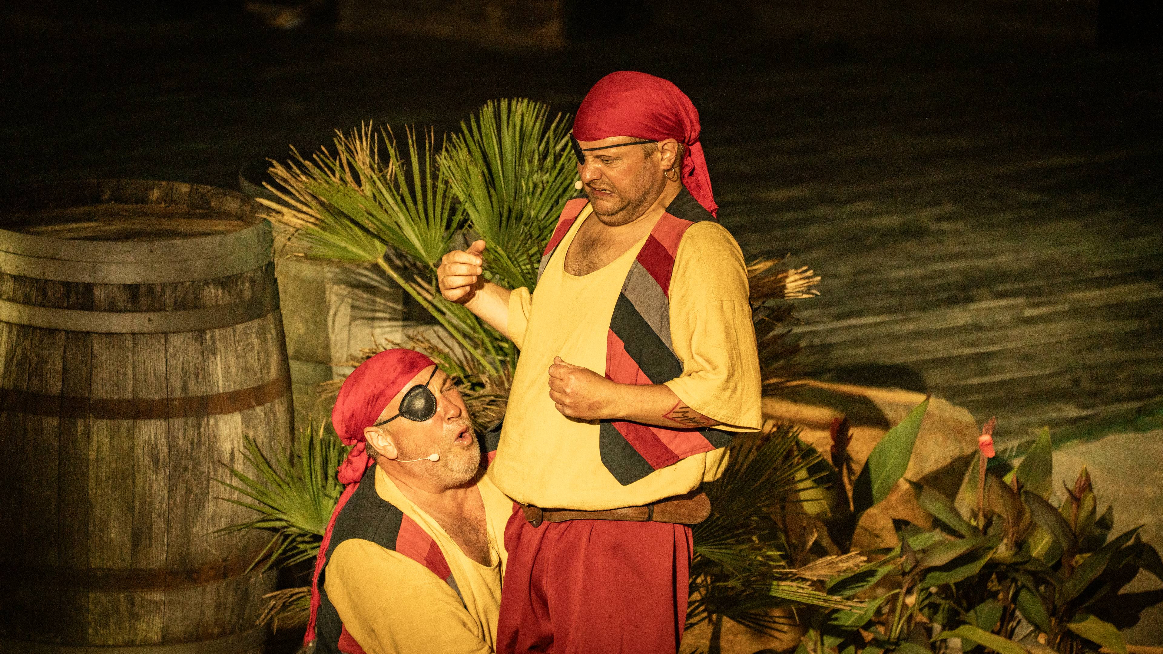 To skuespillere i piratkostymer opptrer på en teaterscene med en tønne og palmer i bakgrunnen. (Bildebeskrivelsen er laget av en KI-tjeneste)