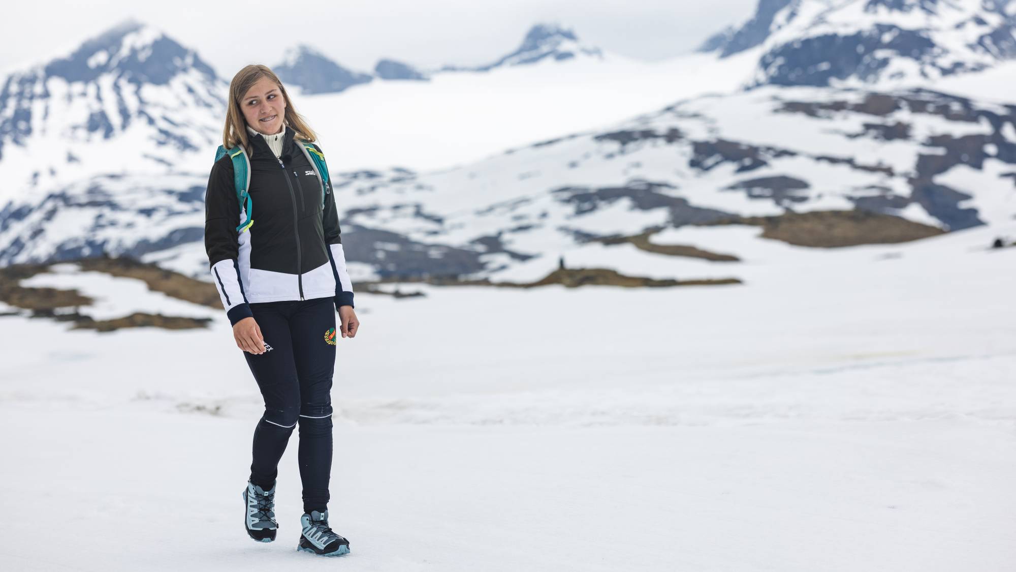 En jente med lyst hår og tannregulering går i et snødekket fjellandskap. Hun har på seg en svart og hvit turjakke, mørke turbukser og en grønn tursekk. Hun smiler forsiktig mens hun ser til siden.