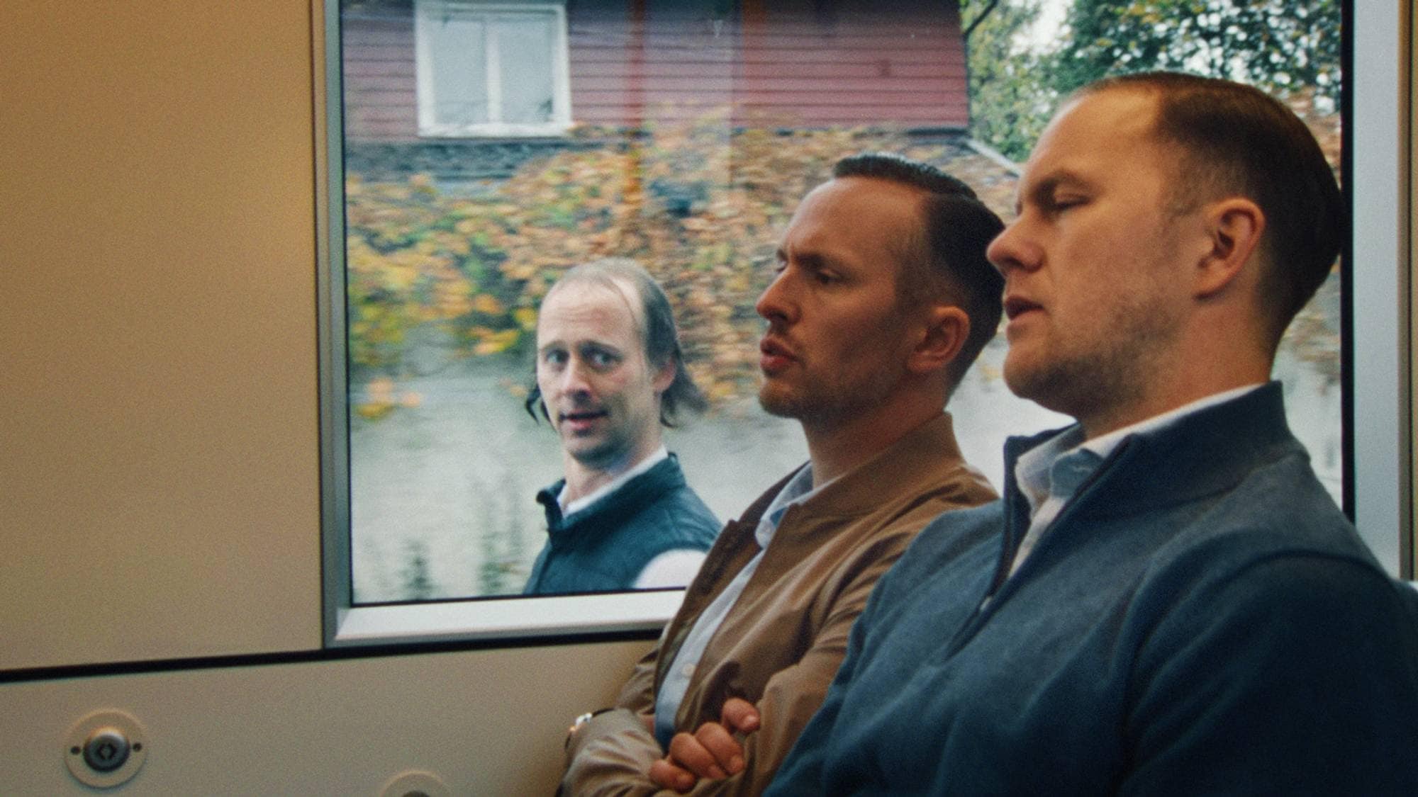 Vi ser tre menn. To av dem sitter i forgrunnen inne på trikken. En mann i bakgrunnen står utenfor vinduet, og løper ved siden av trikken for å holde følge. Utenfor toget er det trær med høstfarger.