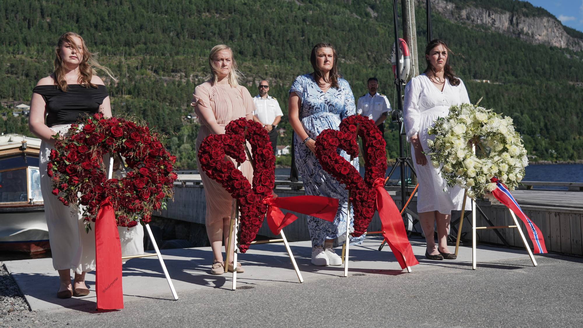 Fire kvinner står ved siden av hver sin blomsterkrans på en brygge ved vannet. (Bildebeskrivelsen er laget av en KI-tjeneste)