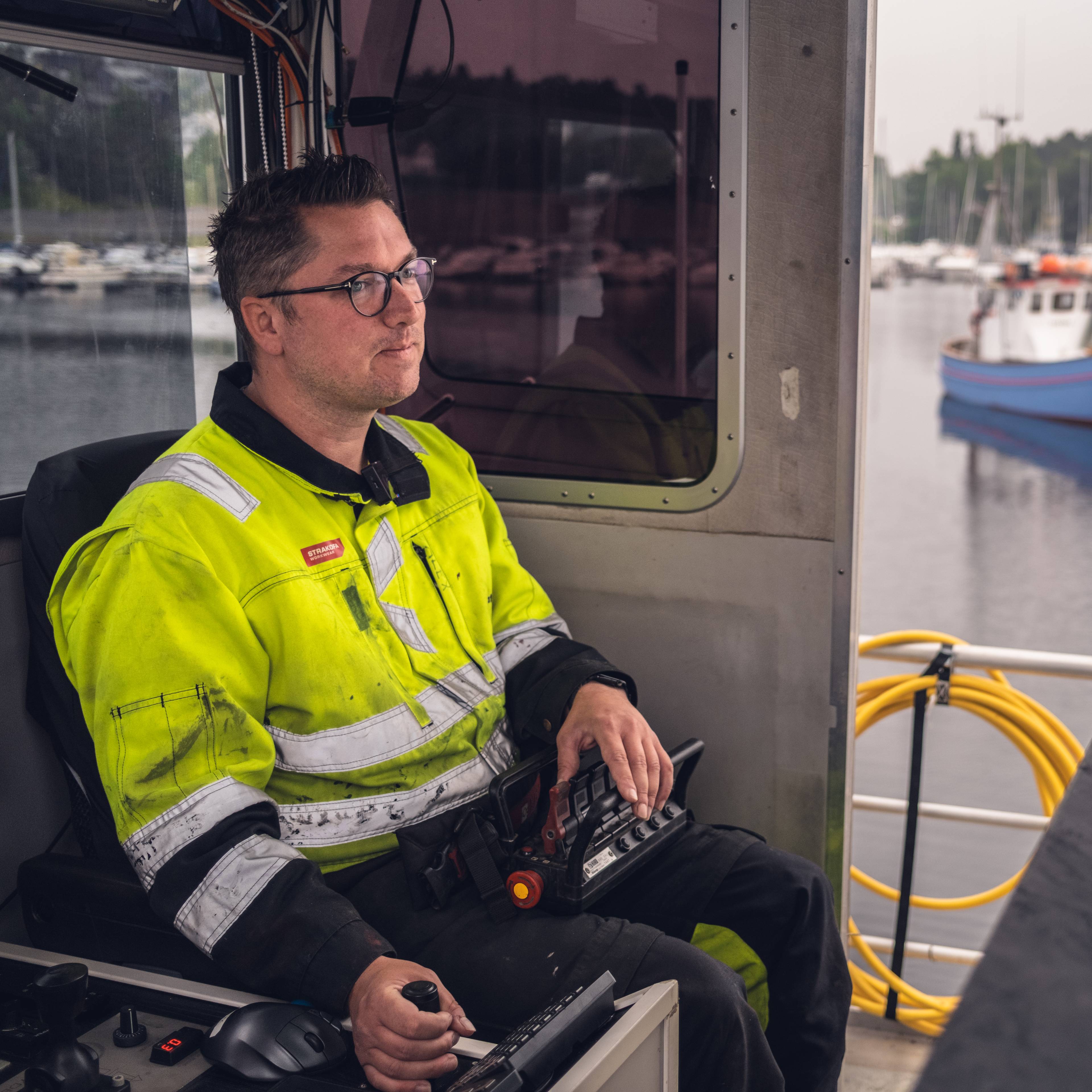 En mann i gul refleksvest sitter i førerhuset på et fartøy. Han holder en fjernkontroll i hendene og ser ut av vinduet. Utenfor ligger flere båter til ankers. (Bildebeskrivelsen er laget av en KI-tjeneste)
