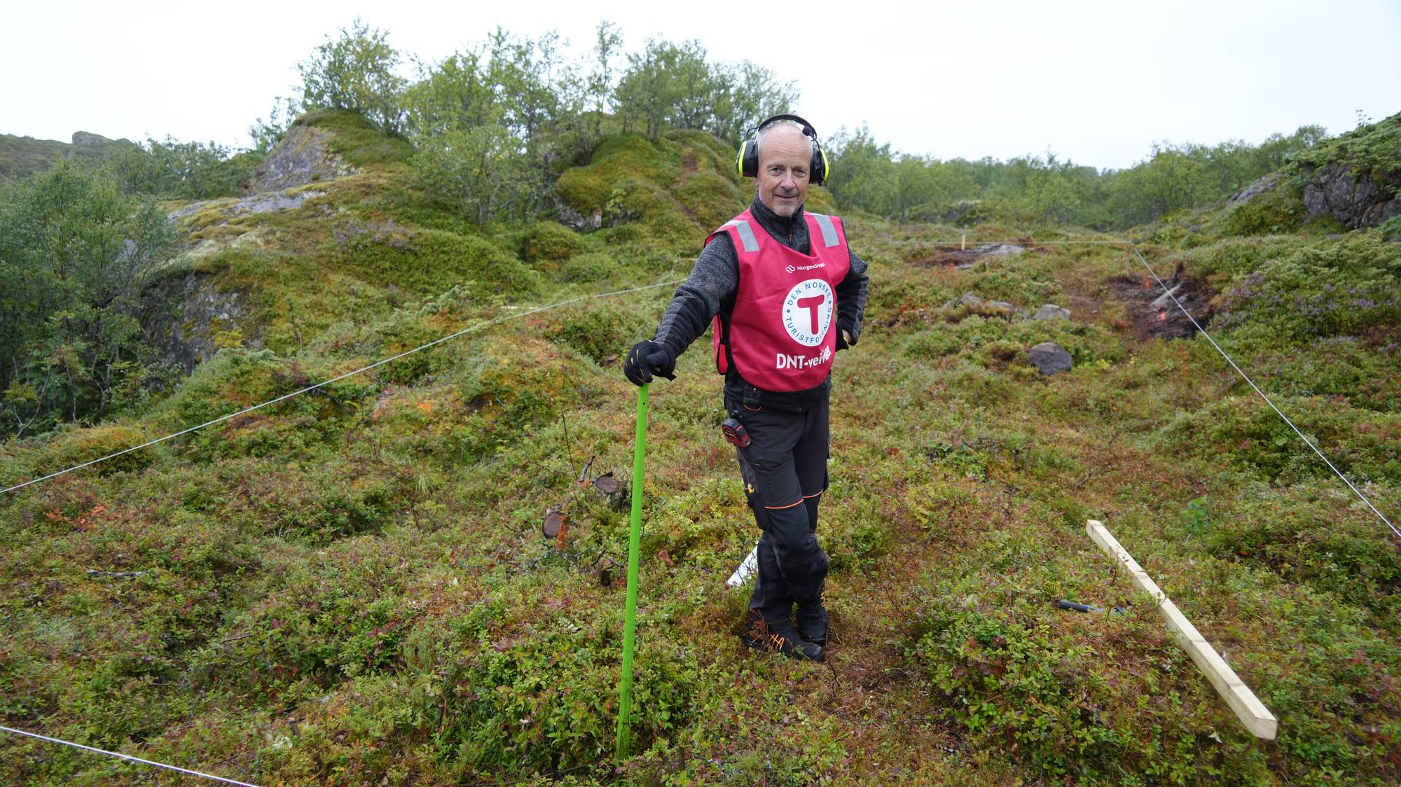 Mann i DNT-vest står i et terreng med en merkestav, omgitt av snor markert for sti. (Bildebeskrivelsen er laget av en KI-tjeneste)