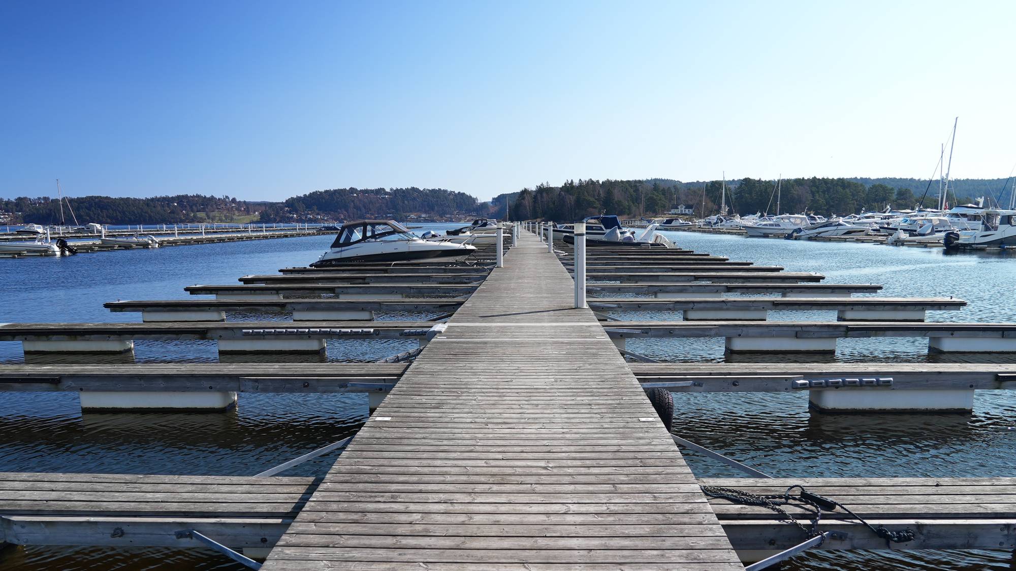 En trebrygge strekker seg mot et ettermiddagssol på en marin. Flere tomme båtplasser ligger langs bryggen. (Bildebeskrivelsen er laget av en KI-tjeneste)