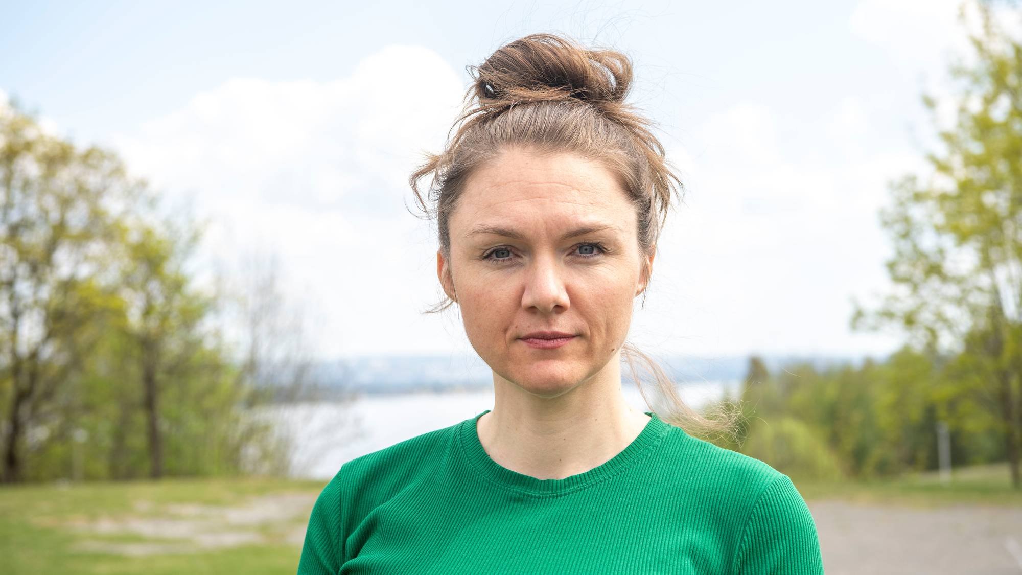 Kvinne med håret i en toppknute ser mot kamera. Hun har på seg en grønn genser. I bakgrunnen er det trær og vann. (Bildebeskrivelsen er laget av en KI-tjeneste)