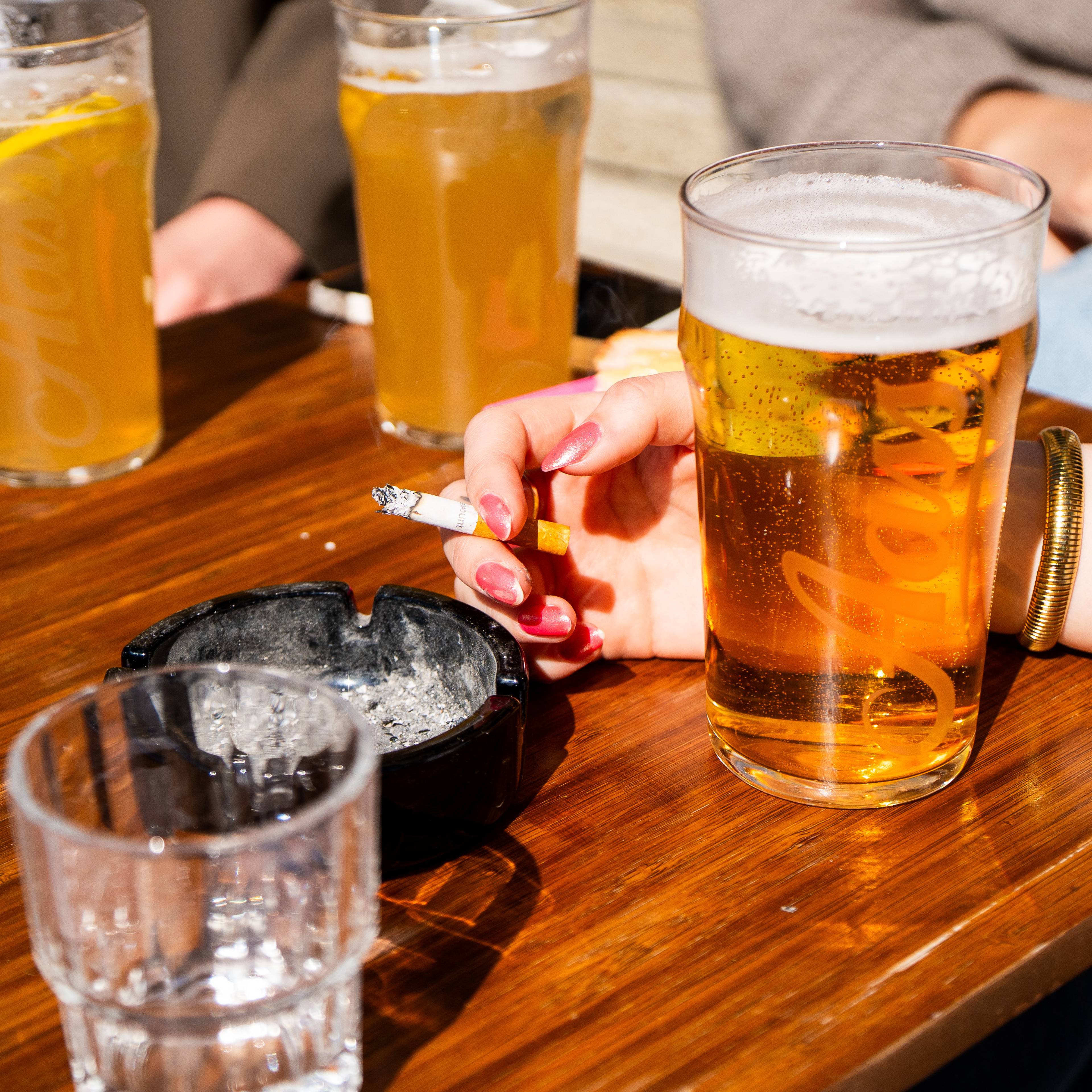 Ei kvinne røyker ein sigarett ved et utandørs bord. På bordet står fleire glas med øl og et oskebeger. Kvinna har rosa negler og et armband. Bildet viser ein gruppe menneske som sit rundt et trebord. (Bildebeskrivelsen er laget av ein KI-teneste)
