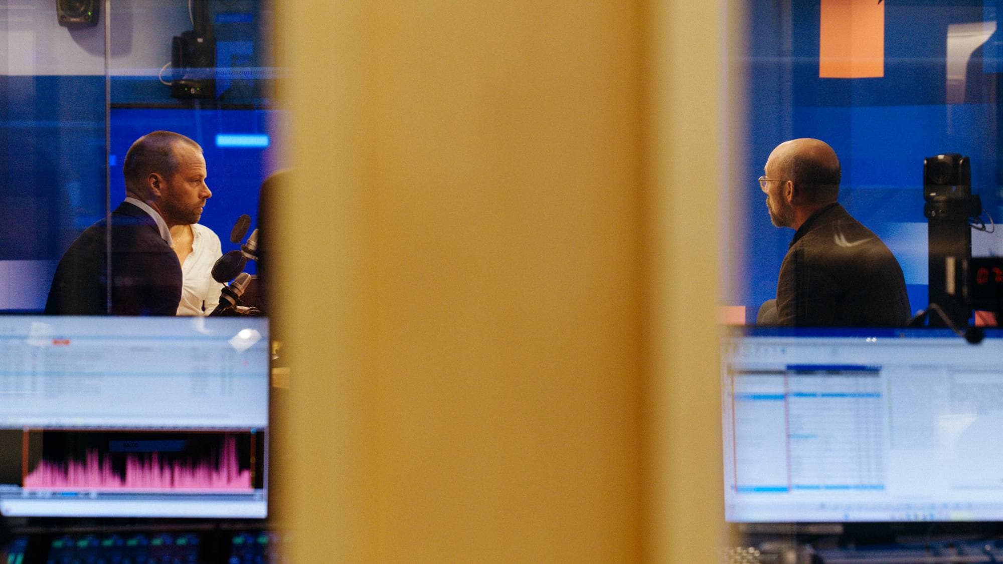 To menn i et radiostudio sett gjennom et vindu, med skjermer og miksepult i forgrunnen. 