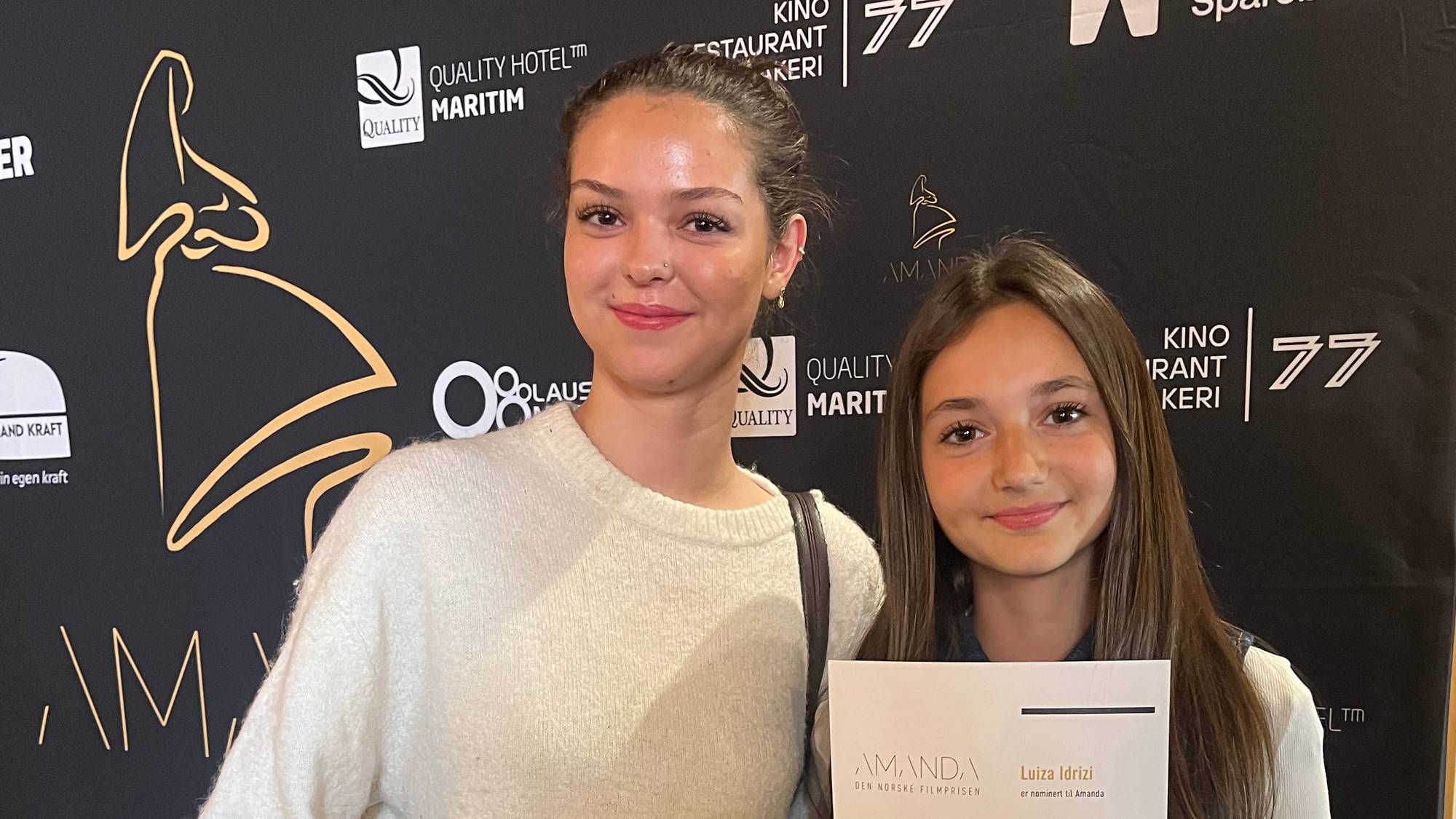 To kvinner holder diplomer fra Amandaprisen. Kvinnen til venstre er Anna Drakopoulou Sverrisdóttir, og kvinnen til høyre er Luiza Idrizi. De står foran en svart bakgrunn med sponsorenes logoer. Til høyre i bildet er en brannslukker. (Bildebeskrivelsen er laget av en KI-tjeneste)