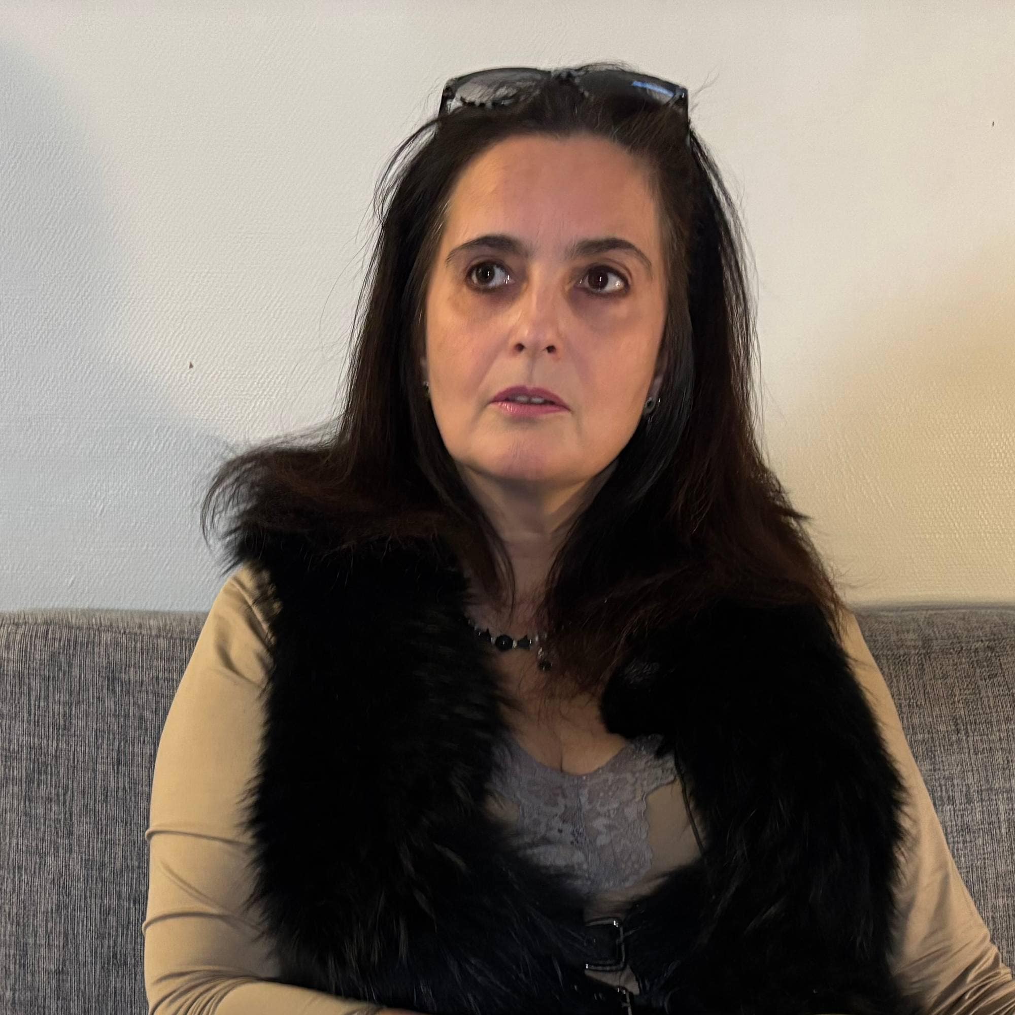 En person sitter avslappet på en grå sofa, iført en beige drakt og en svart jakke. Vedkommende holder en gul kopp med et mønster. Bakgrunnen er en lys vegg, som gir en enkel og nøytral kontekst. Omgivelsene virker innbydende og hjemmekoselige.