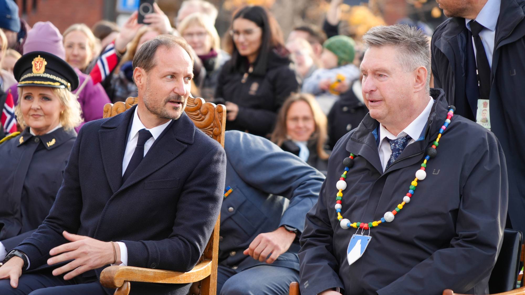 Kong Haakon og ordfører Rune Edvardsen i Narvik sitter på trestoler og snakker med hverandre på torget i Narvik. Mange narvikinger sitter bak og venter på programmet på scenen.