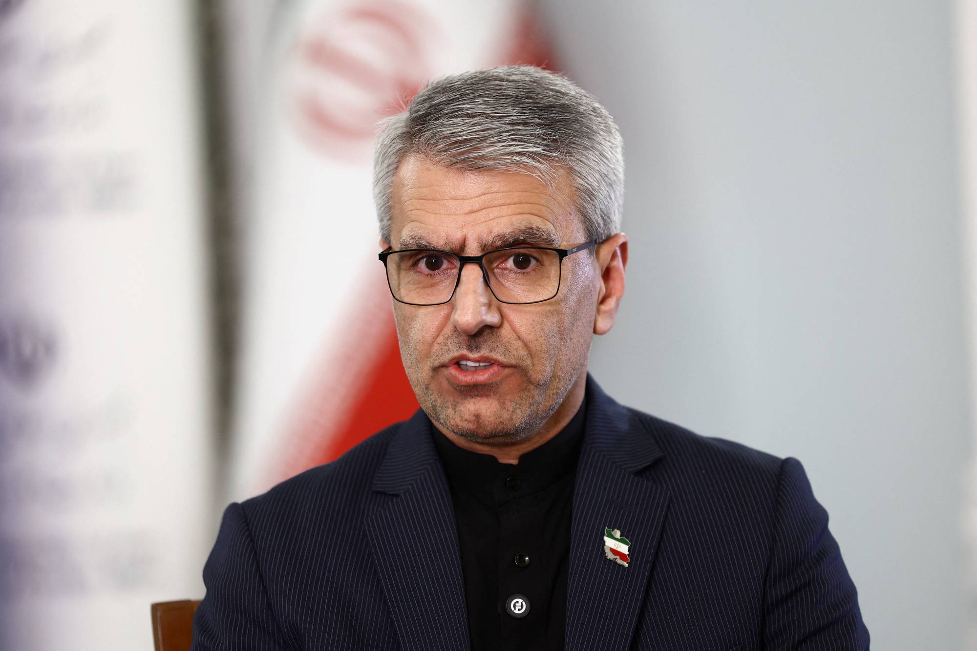 A man in glasses speaks in front of a blurred background. He wears a dark suit and a black shirt. There is a small badge on his lapel depicting the flag of Iran. The image has a watermark. (Bildebeskrivelsen er laget av en KI-tjeneste)
