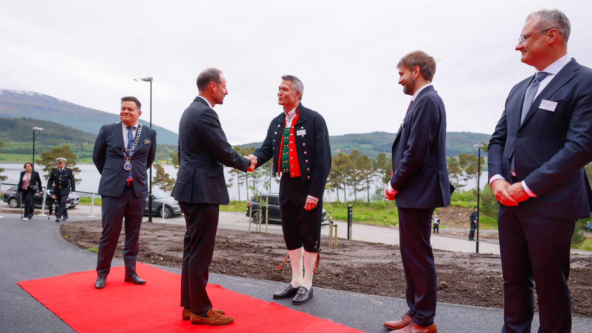 To menn i dress hilser på hverandre på en rød løper. En av mennene har på seg bunad. I bakgrunnen sees et vann og fjell. (Bildebeskrivelsen er laget av en KI-tjeneste)