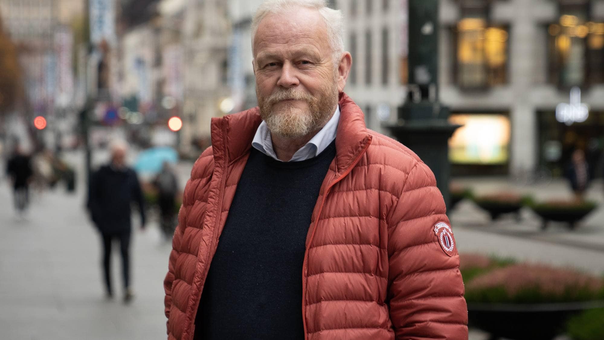 En mann med rød jakke ser i kamera. I bakgrunn en bygate. 