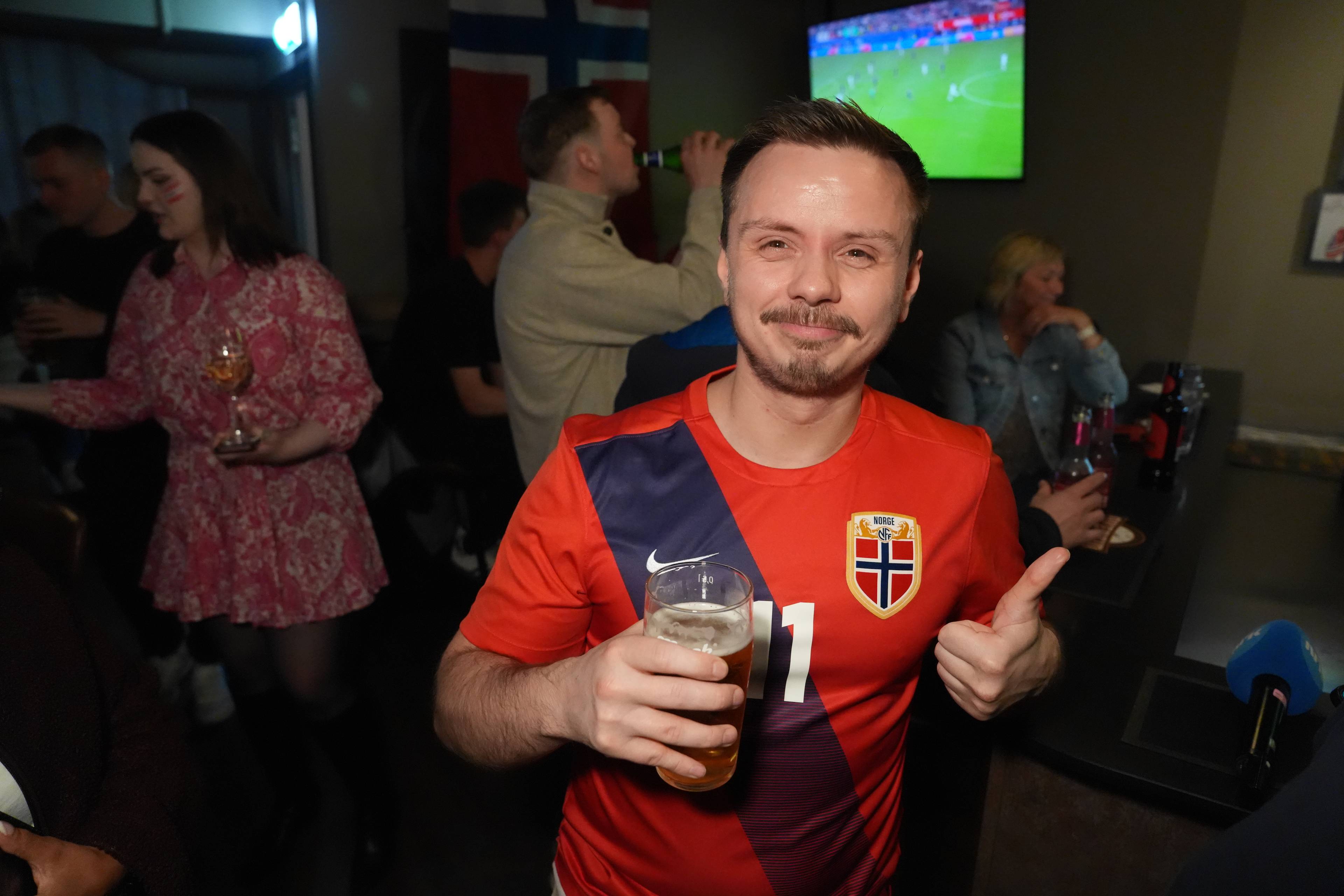 En mann i en norsk fotballtrøye holder et glass øl og gir en tommel opp. I bakgrunnen sitter folk i en bar. En TV viser et fotballkamp.