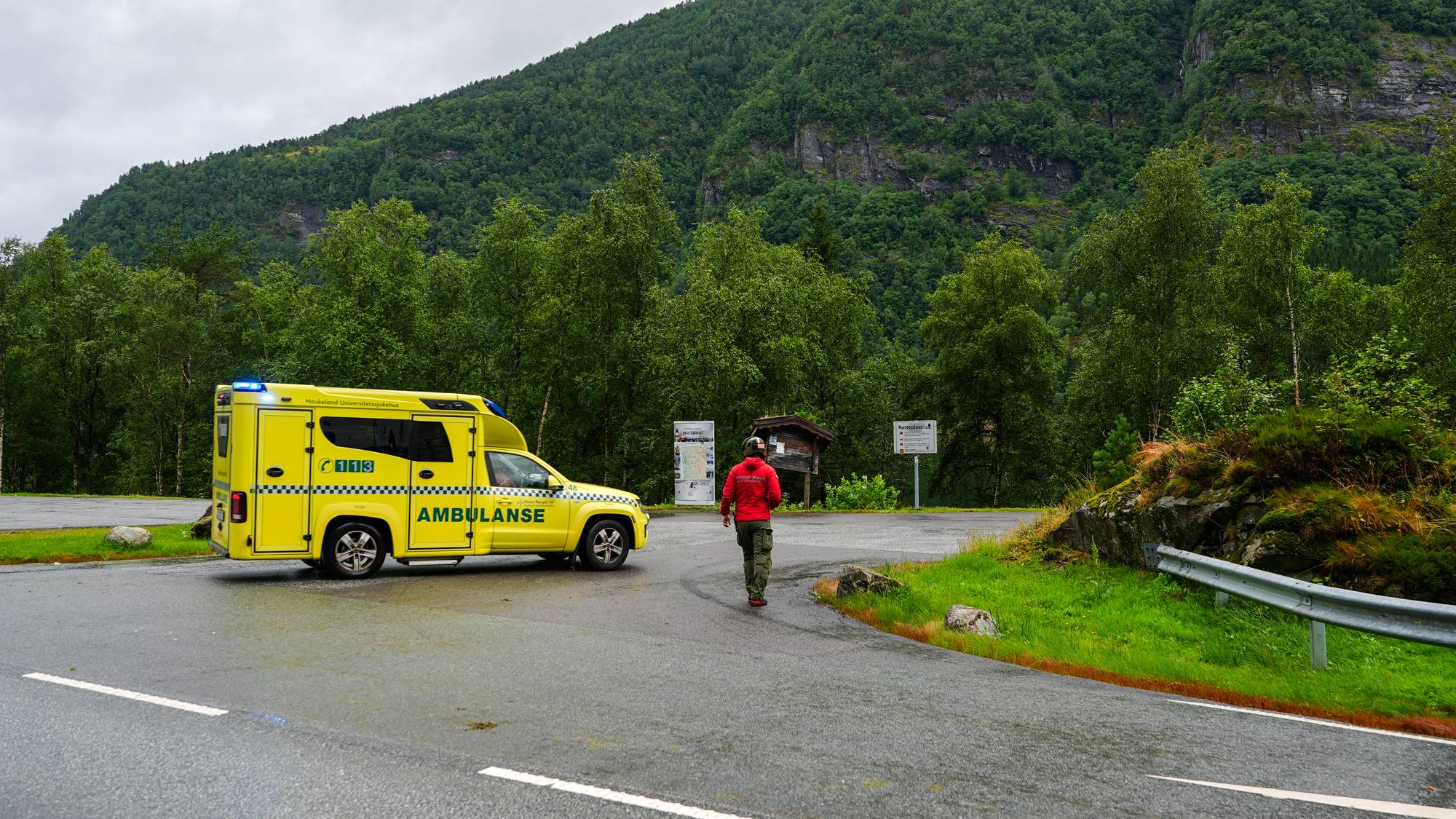 Gul ambulanse står parkert ved en vei i et fjellandskap, med en person i rød jakke gående mot den. 